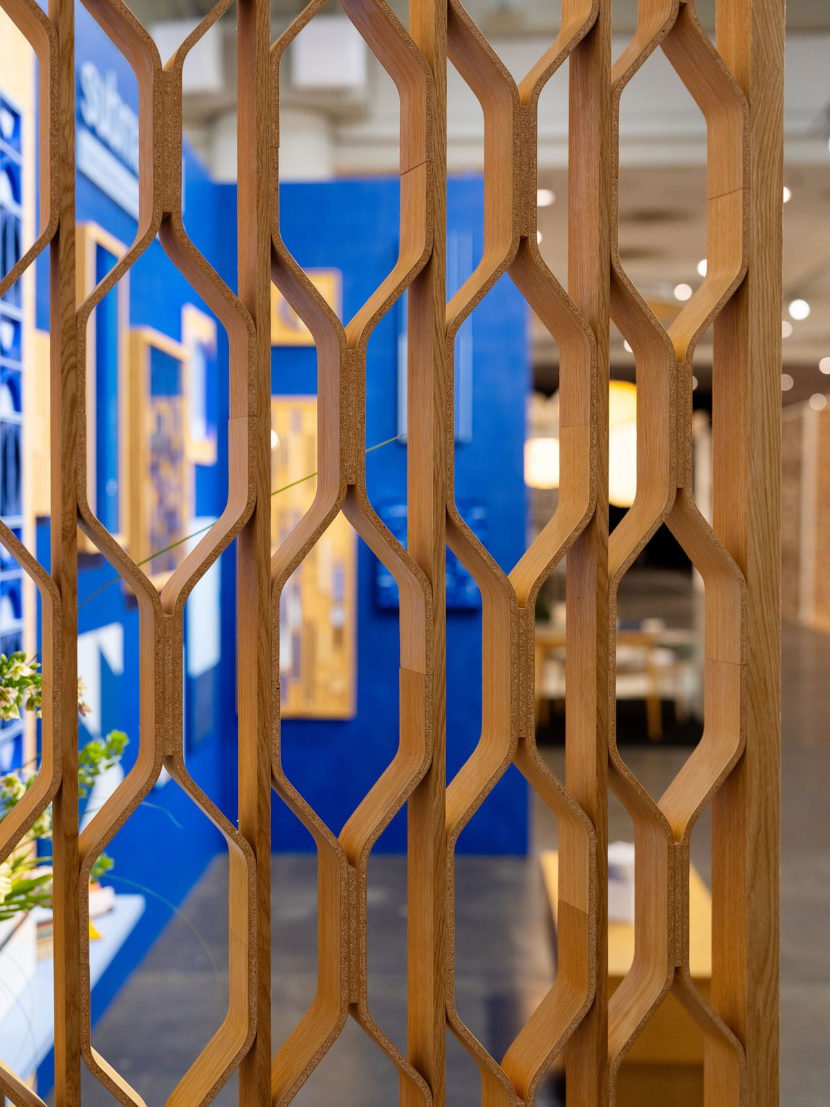 A close-up of a modern wooden geometric lattice screen or partition, with a blurred indoor showroom or office space in the background featuring blue walls and framed displays.