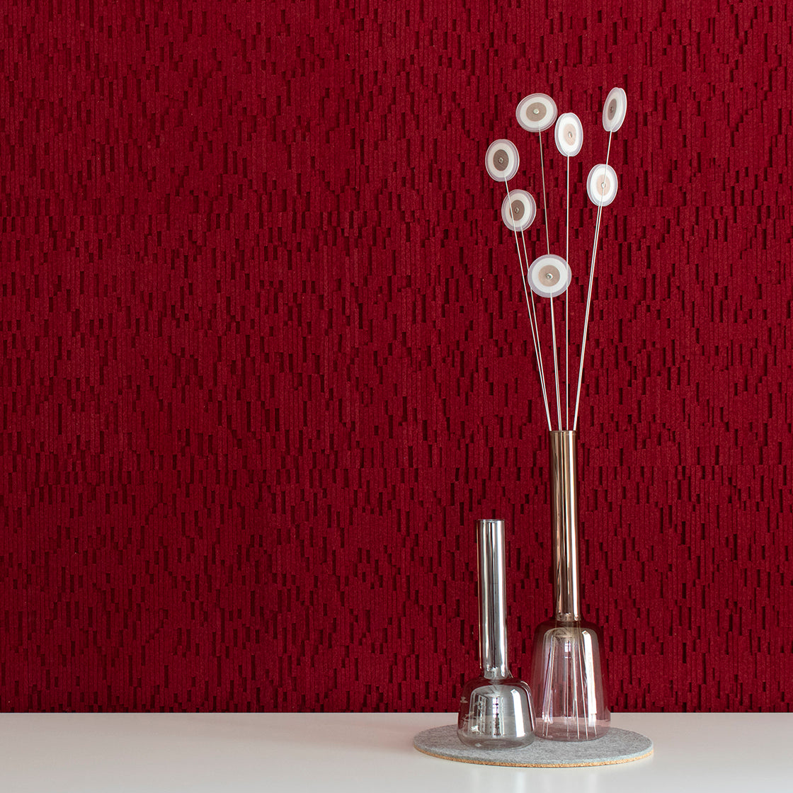 A red wool felt wall covering with narrow vertical cutouts. The surface in front of the wall is white with a gray felt mat. Two tall narrow glass vases and a few abstract flowers.