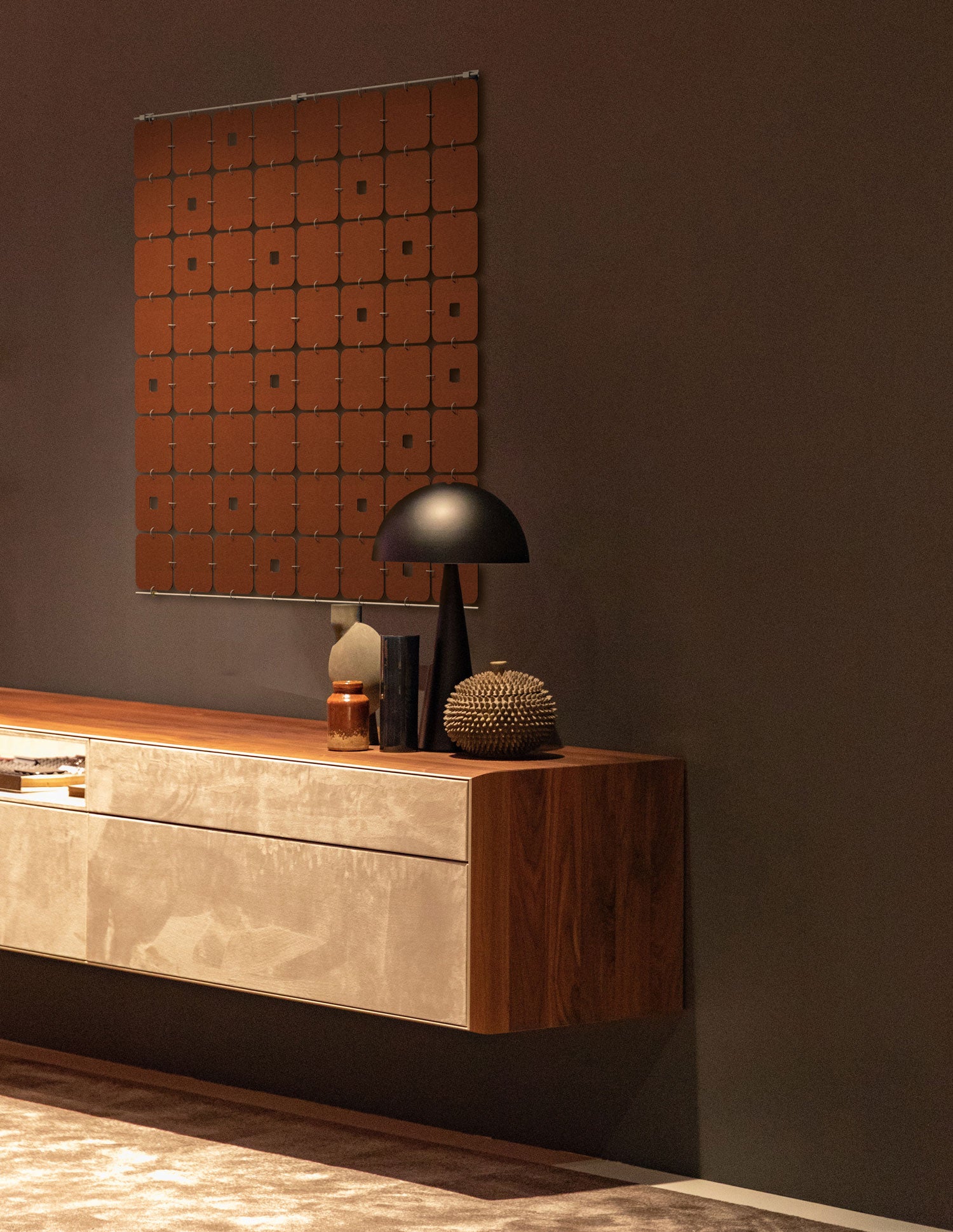 A felt wall hanging of soft squares with occasional square keyholes hangs in a dark room with brown walls and a floating cabinet.