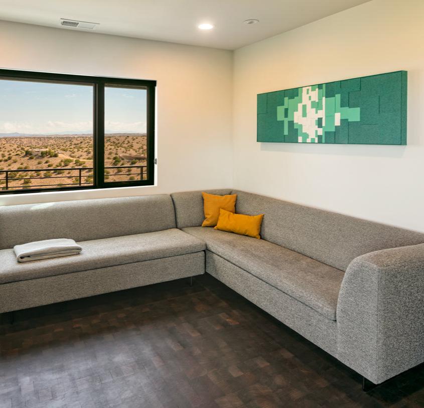 A bright living room with a sleek L-shaped couch. At right a rectangular felt wall panel in blocks of green and whites. To the left a large window showing the scrubby high desert.