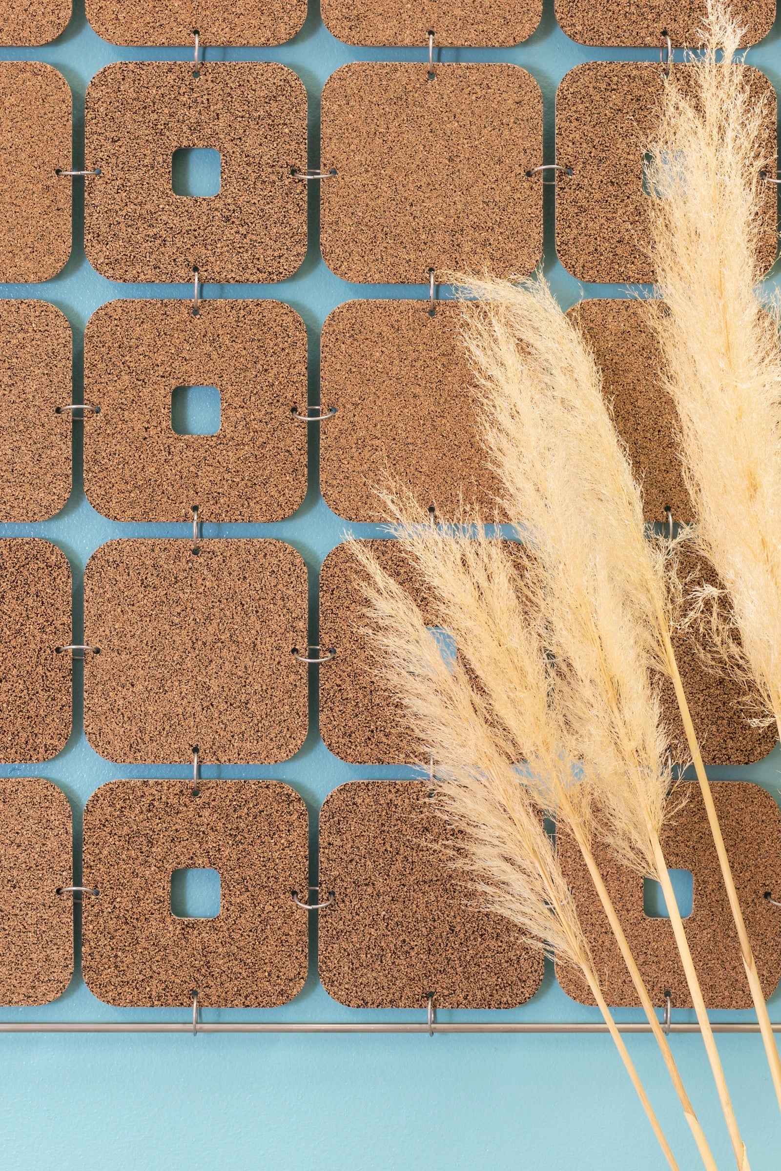 A closeup of a cork wall hanging with softly curved squares and occasional keyholes. A round metal dowel hangs at the bottom and tan feathery stalks are in the forefront.