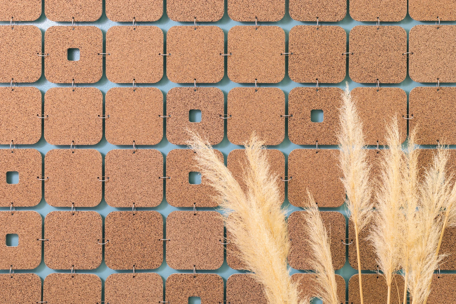 A closeup of a cork wall hanging with softly curved squares and occasional keyholes. Tan feathery stalks are in the forefront.