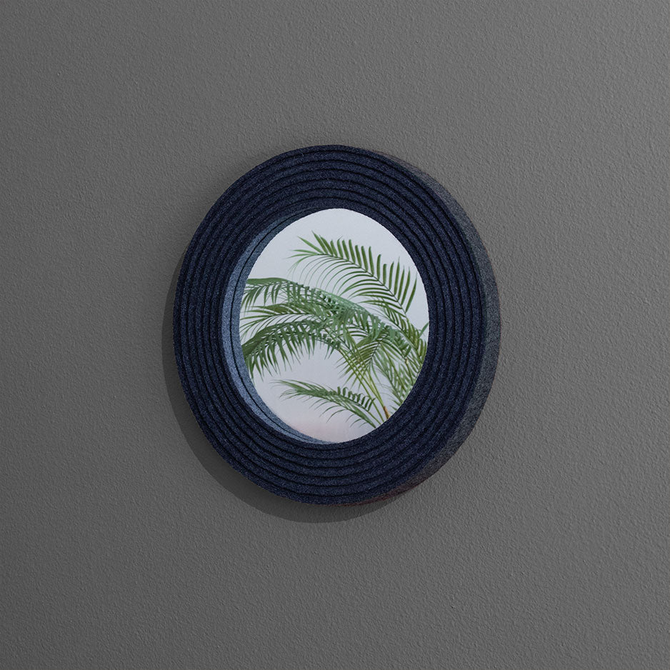 A small round circle with a gray heathered felt frame. It hangs on a gray wall and reflects a firm in the mirror.