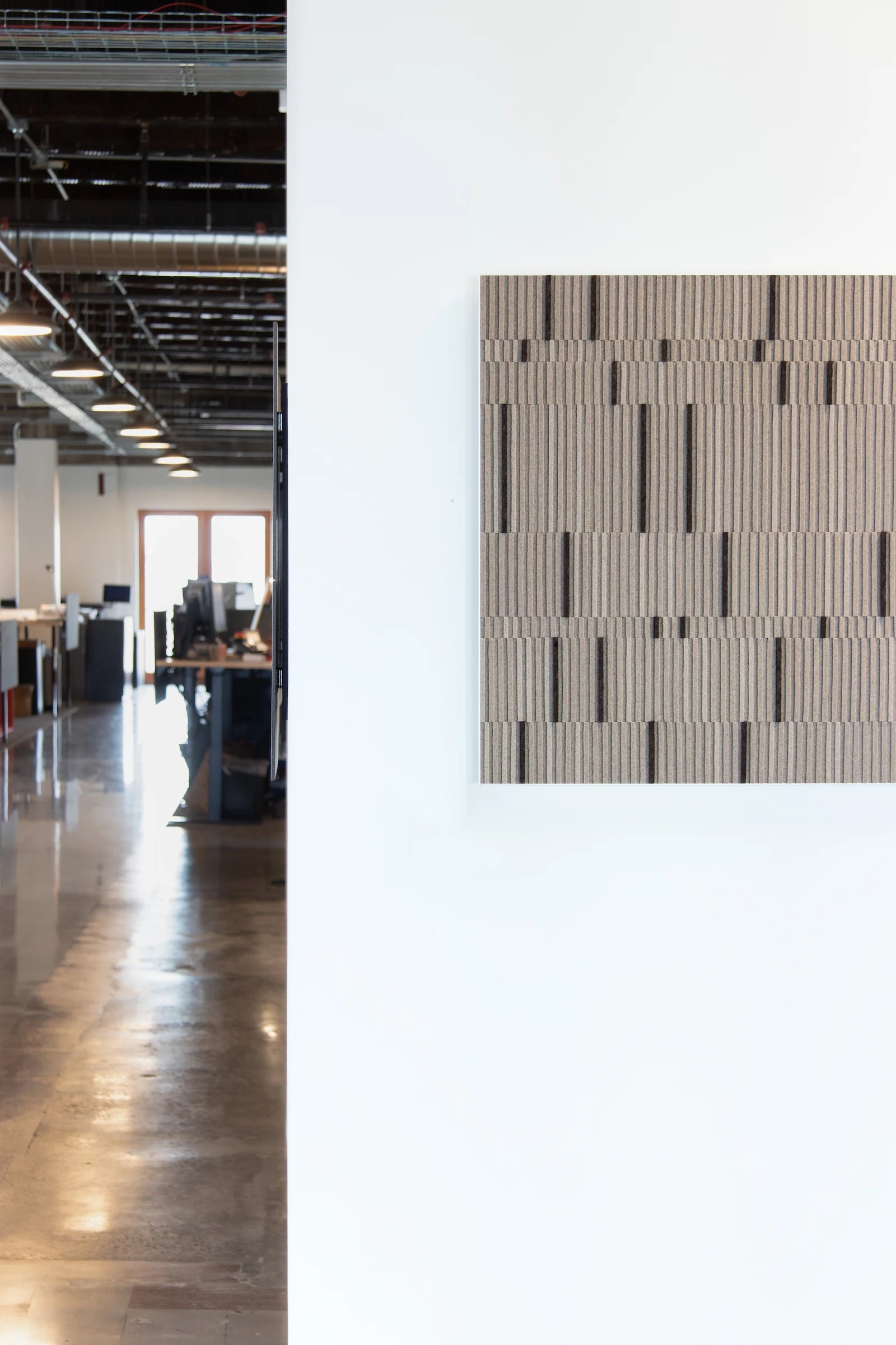 Circadian No. 1 wall art piece of tan rows of alternating height felt with random bands of smoked cork in a square white metal cradle. It is hung on a while wall in n office setting with desks and computers in the distance.