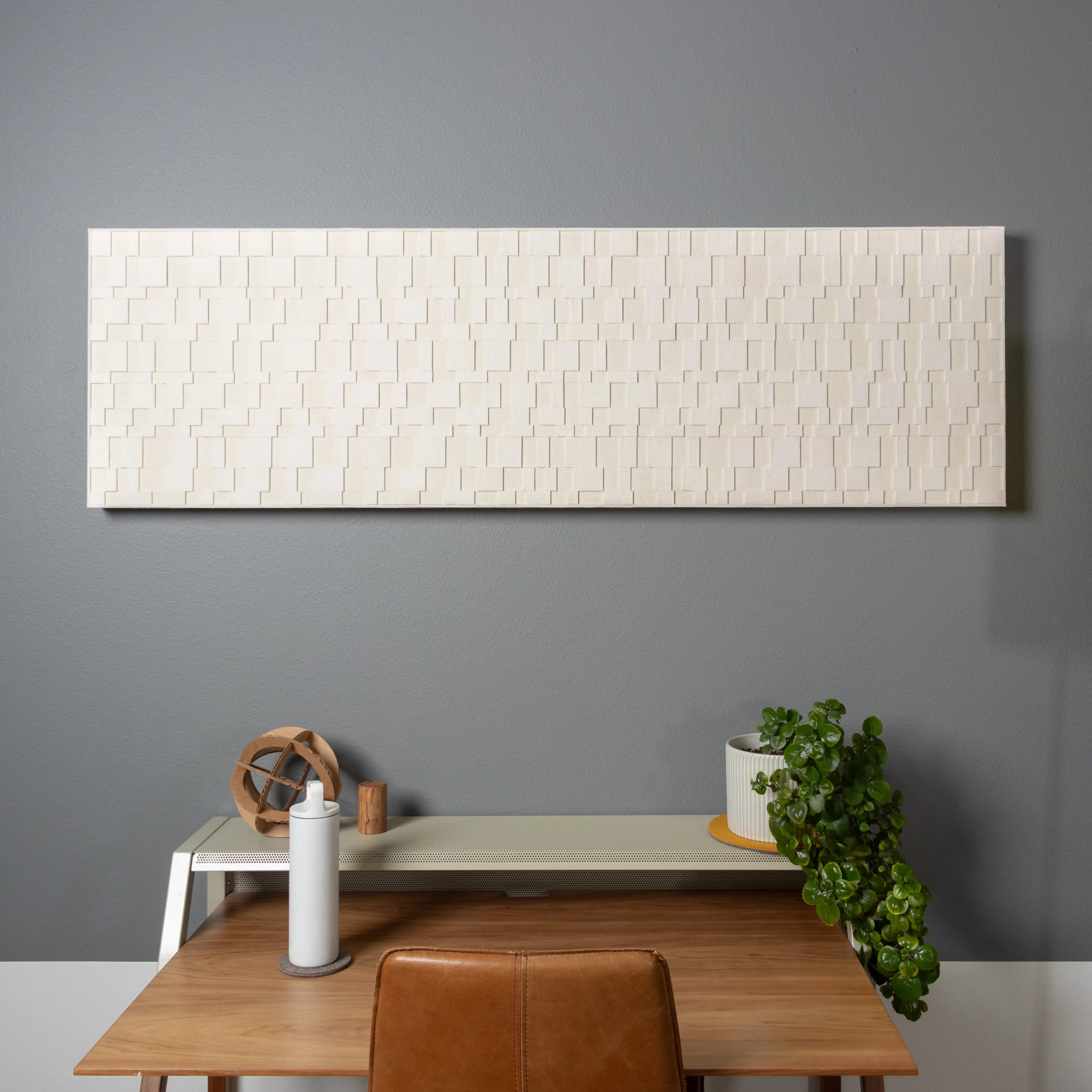 An Aerial wall panel. A rectangular wall art piece of white felt in various sizes and layers. It hangs on a gray wall over a modern desk with a plant, water bottle and other knick knacks.