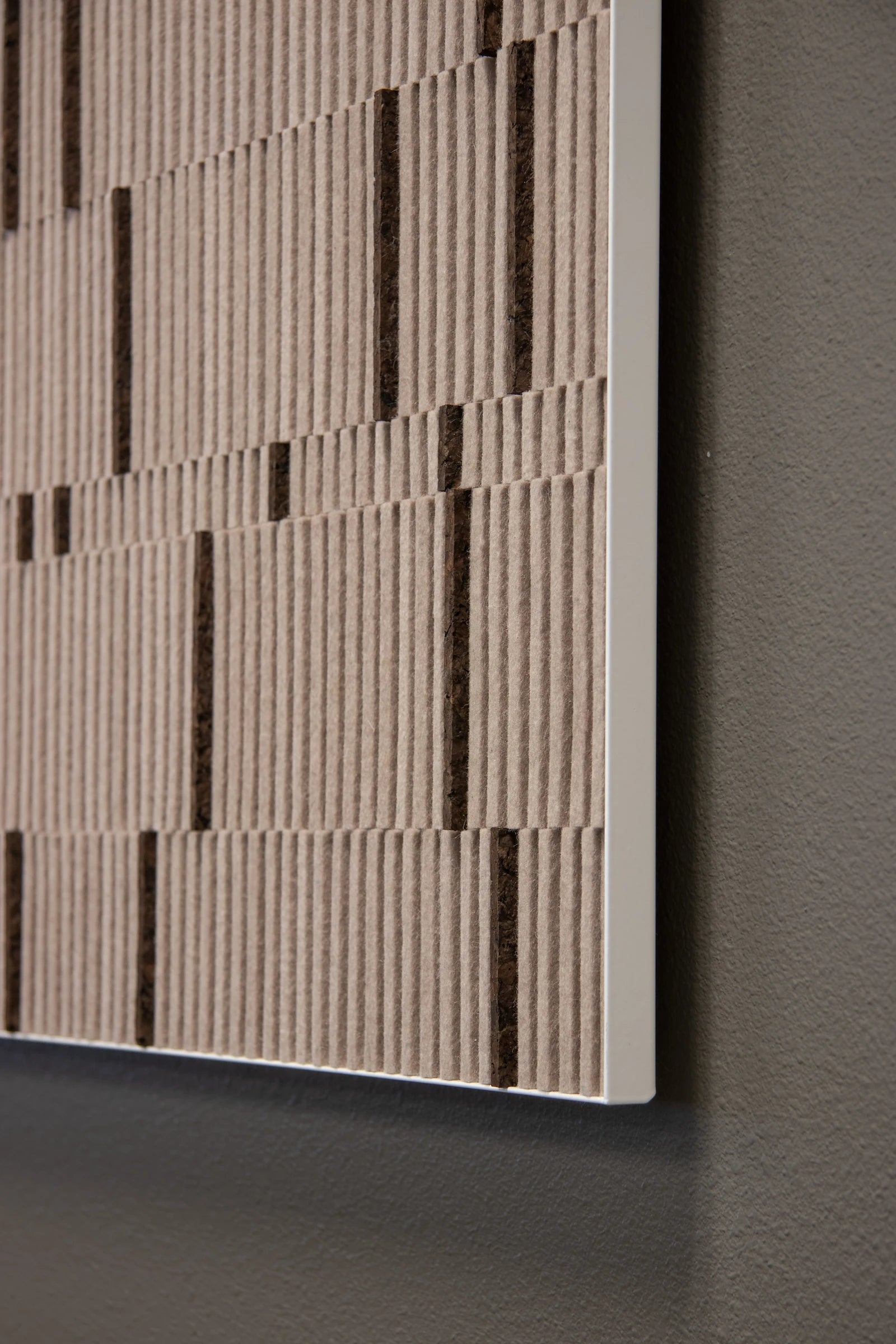 Circadian No. 1 wall art piece of tan rows of alternating height felt with random bands of smoked cork in a square white metal cradle seen from the corner on a dark gray wall.
