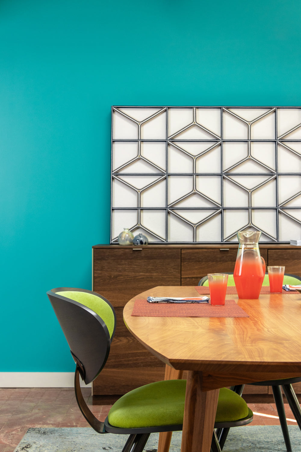 A large rectangular felt wall art panel sits on a sideboard. It is white with a grid and Y-shaped in white and blue felt. The back wall is a bright turquoise. A dining table with green chairs and orange glassware is in the foreground.