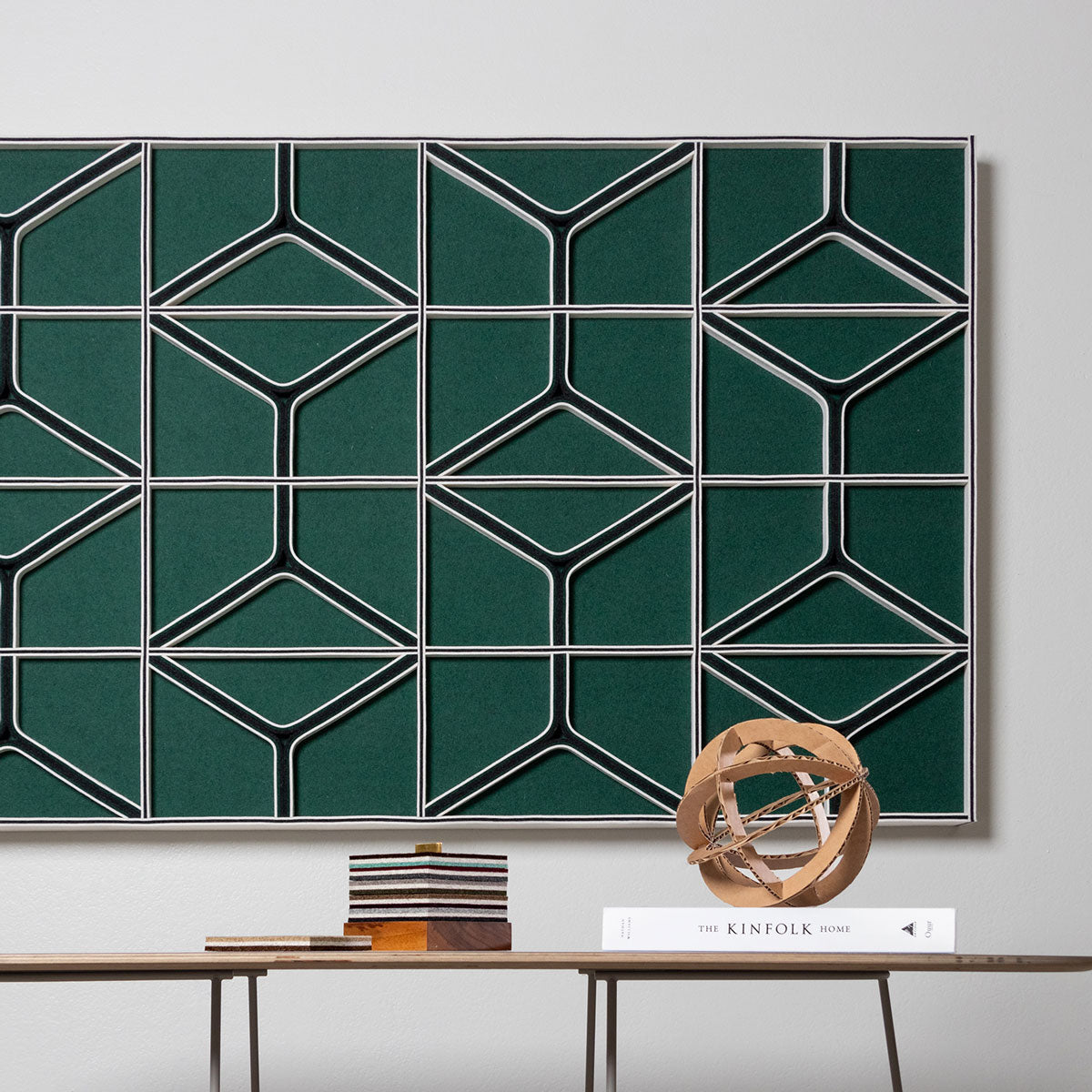 A large felt wall panel with a green face and green and white lines and angles hangs behind a small table with knick knacks.