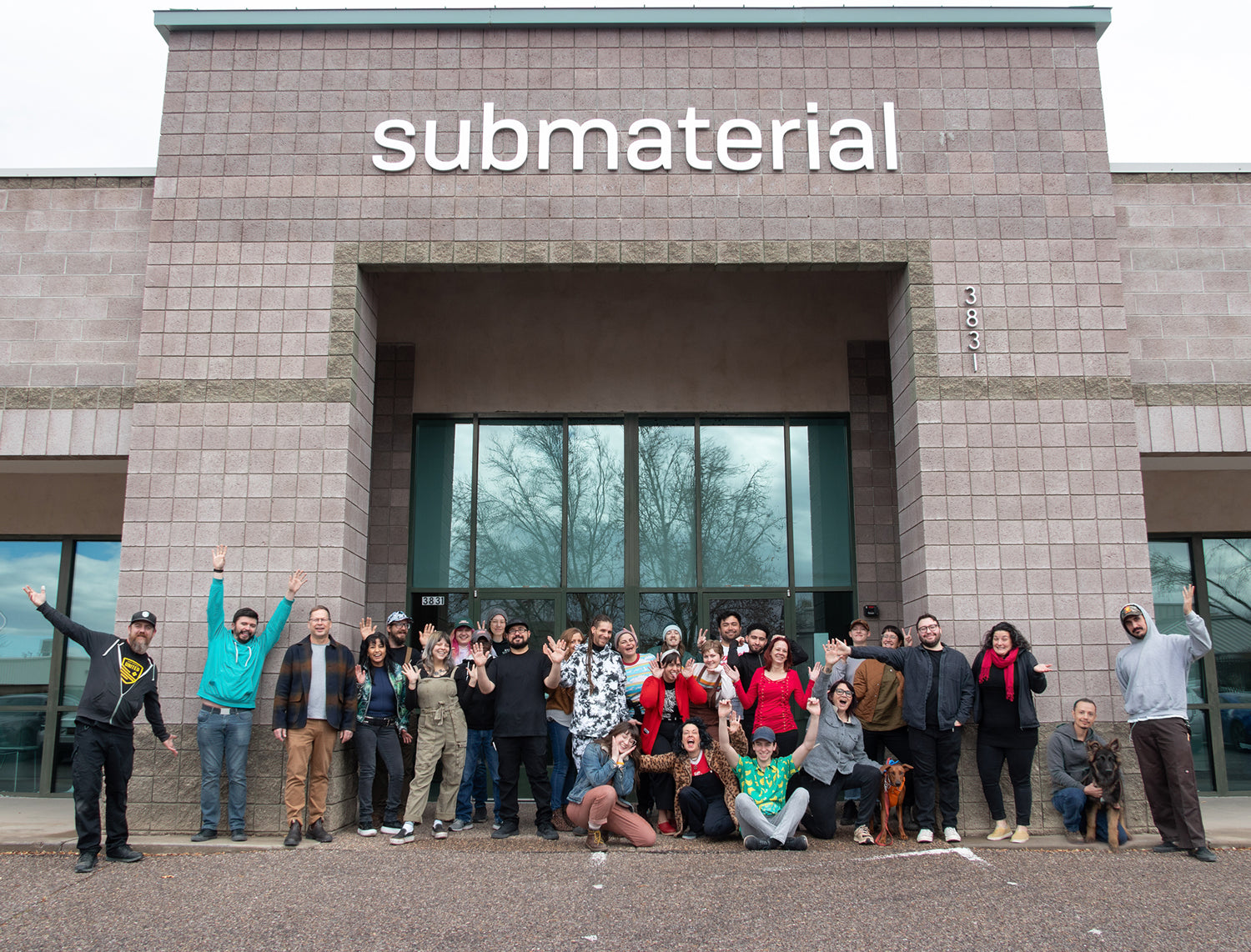A groups of people outside of a building making silly faces and lifting their arms. The building has the word 'submaterial' on the facade.