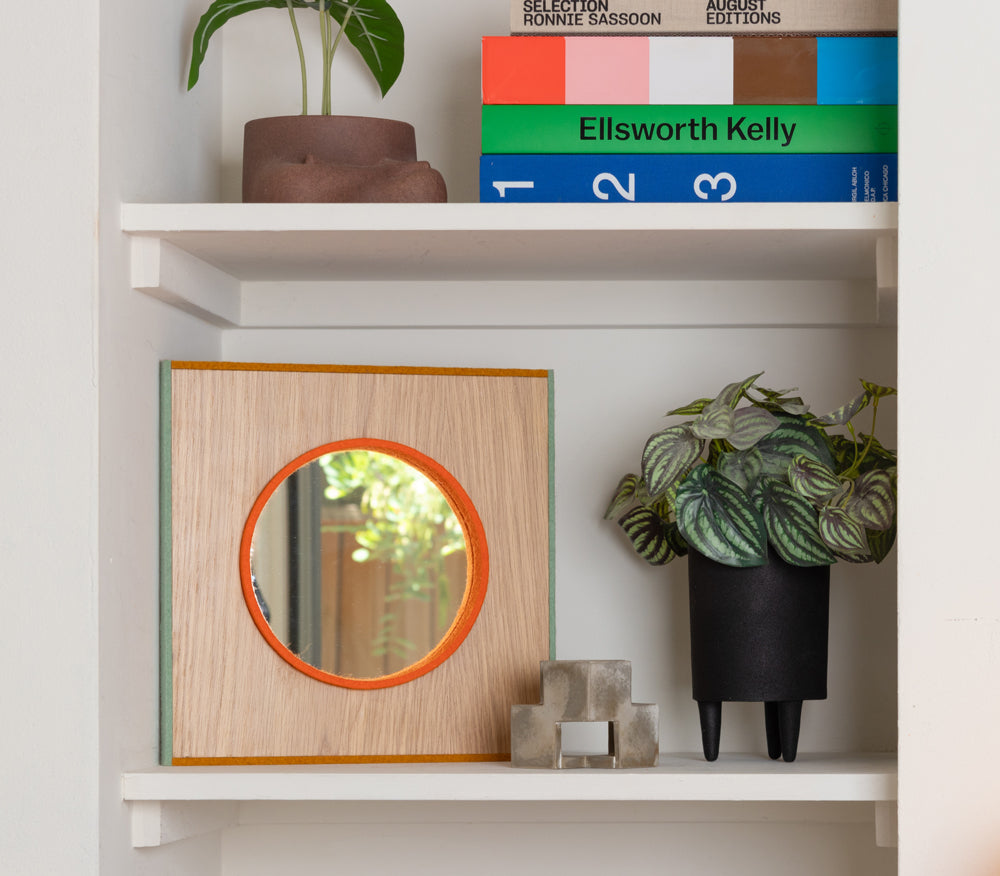 A shelf area with books and plant on the top shelf and a plant and mirror on the shelf below. The small mirror is framed with a strip of orange felt, surrounded by wood surface and the whole thing framed with sage and yellow felt on the edges.
