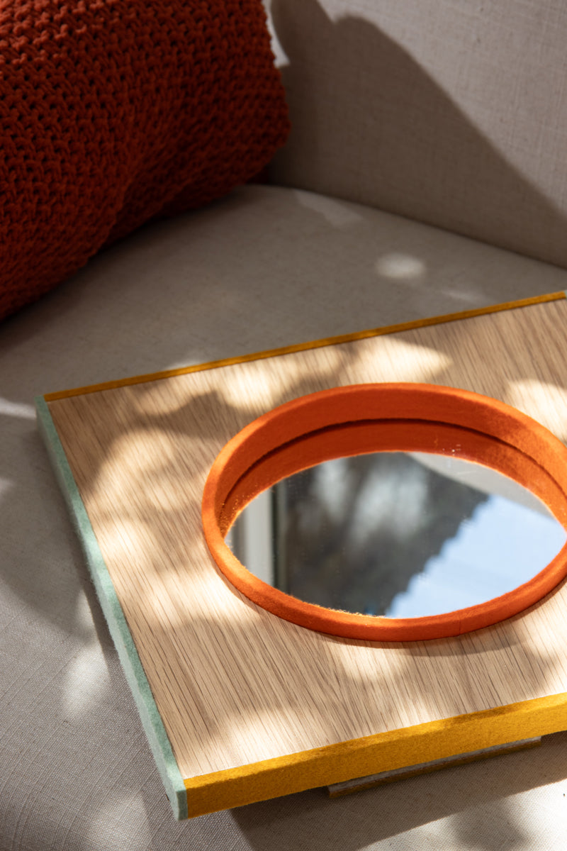 A small mirror framed with a strip of orange felt, surrounded by wood surface and the whole thing framed with sage and yellow felt on the edges. The mirror lays on a couch with dappled sunlight.