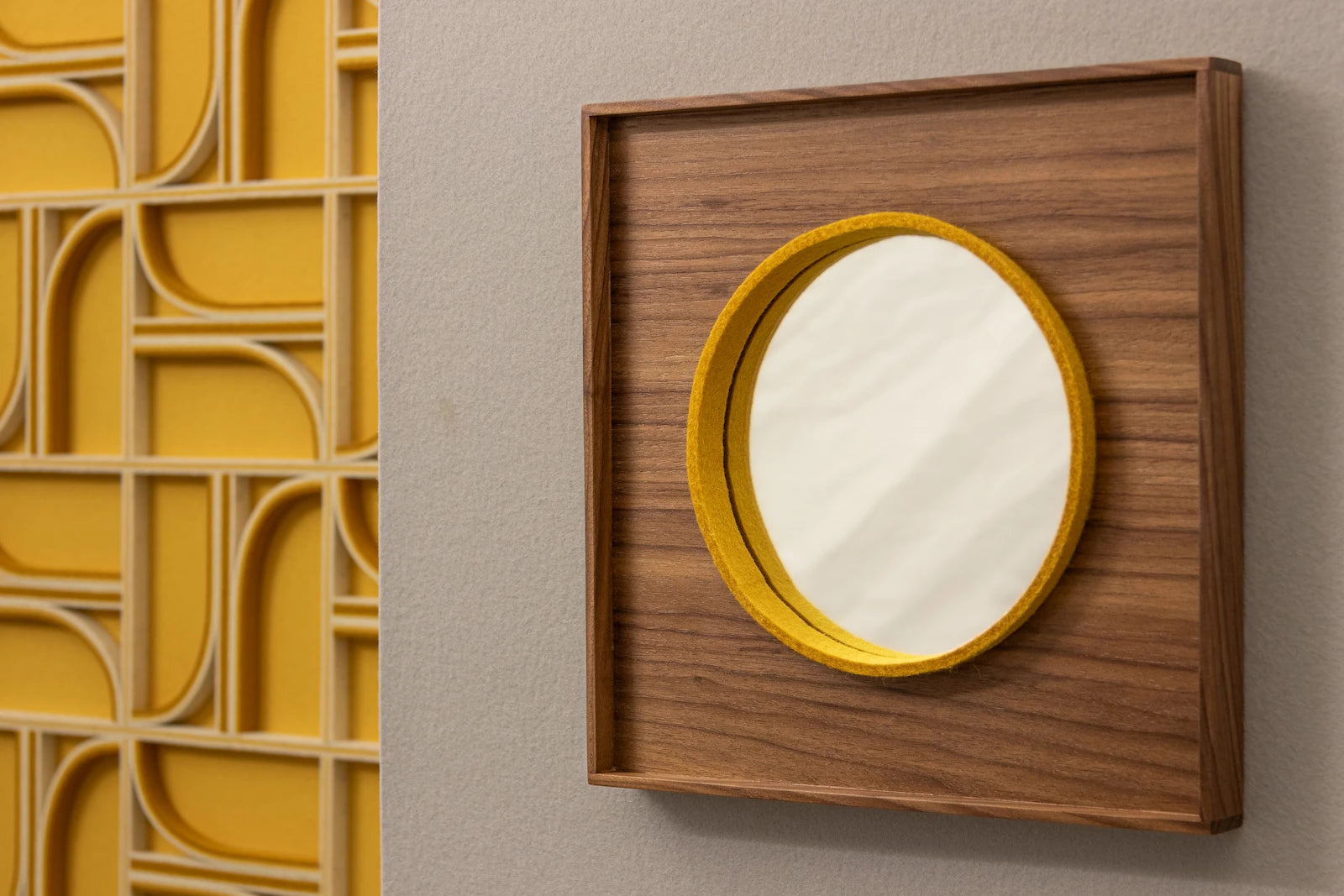 A mirror with a wooden frame and face and a yellow on-edge piece of felt that frames a small circle mirror hangs on a white wall. In the background at left is a white and white felt wall art piece with straight and curved lines.
