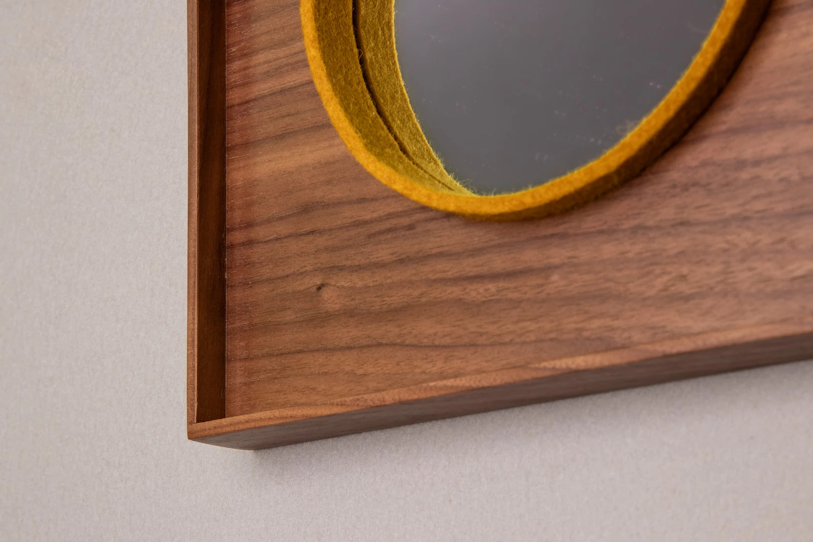 A mirror with a wooden frame and face and a yellow on-edge piece of felt that frames a small circle mirror hangs on a white wall.