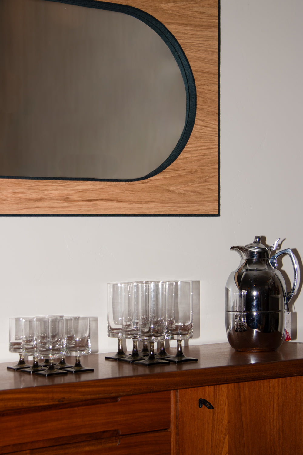 A sideboard with a metal pitcher and glassware site below a large mirror. The mirror is rectangular with a blue frame, wood surface and a pill-shaped reflective surface framed with blue felt.