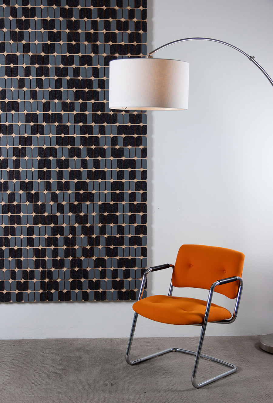 A large felt wall panel of gray and slate blue curved shapes hangs on a white wall. A bight orange cantilevered chair and an arc lamp with white lamp shade can be seen at right.