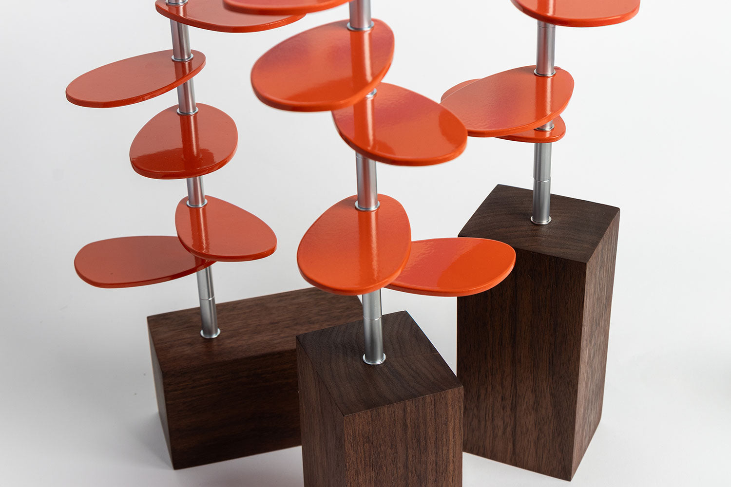 A side/top view of Murmur Trees. Three sculptural objects with walnut wood block bases, brushed metal stems and orange powder-coated oval leaf shapes.