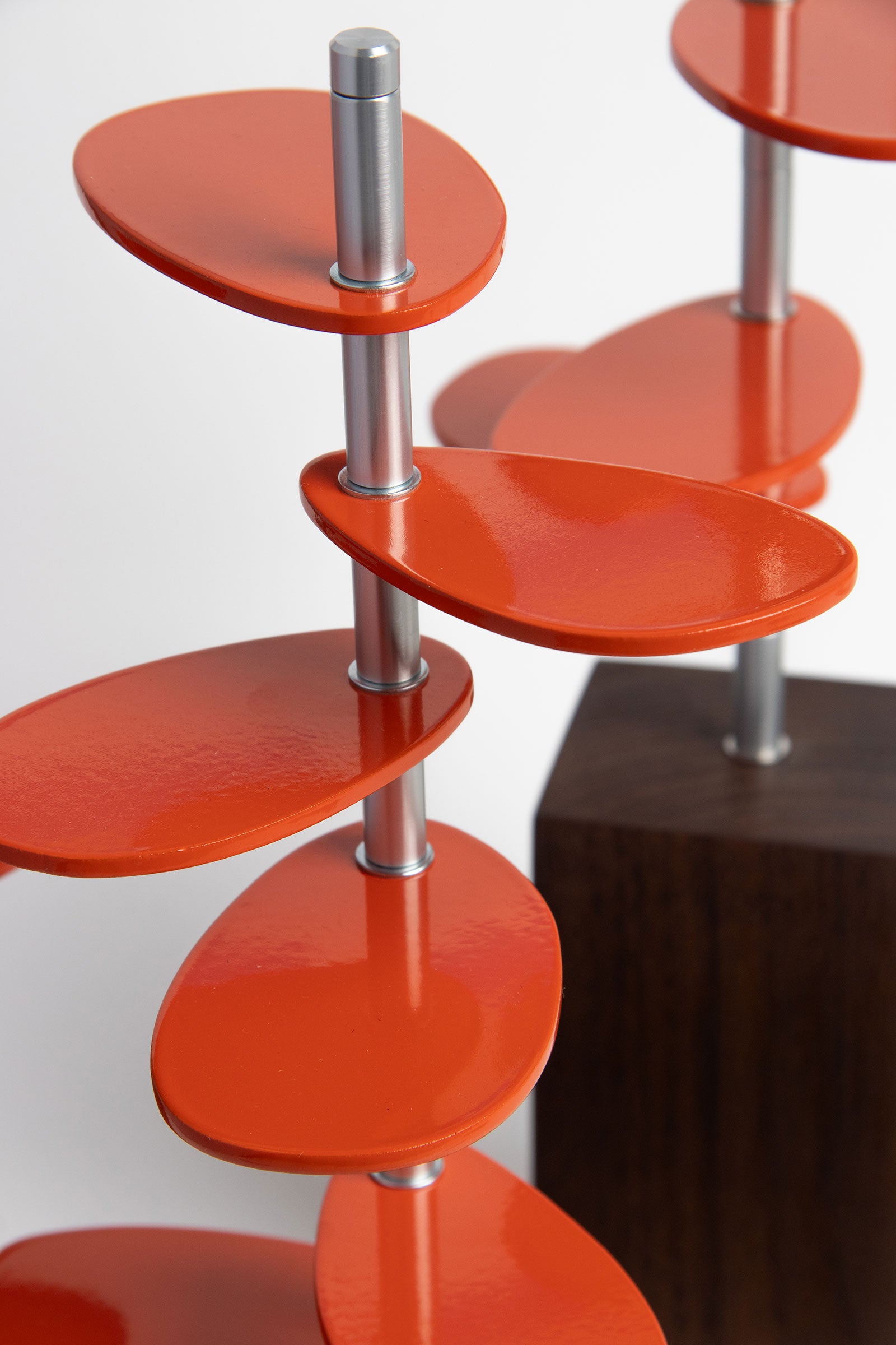 A closeup of Murmur Trees, sculptural objects with walnut wood block bases, brushed metal stems and orange powder-coated oval leaf shapes. #color_Orange metal leaves