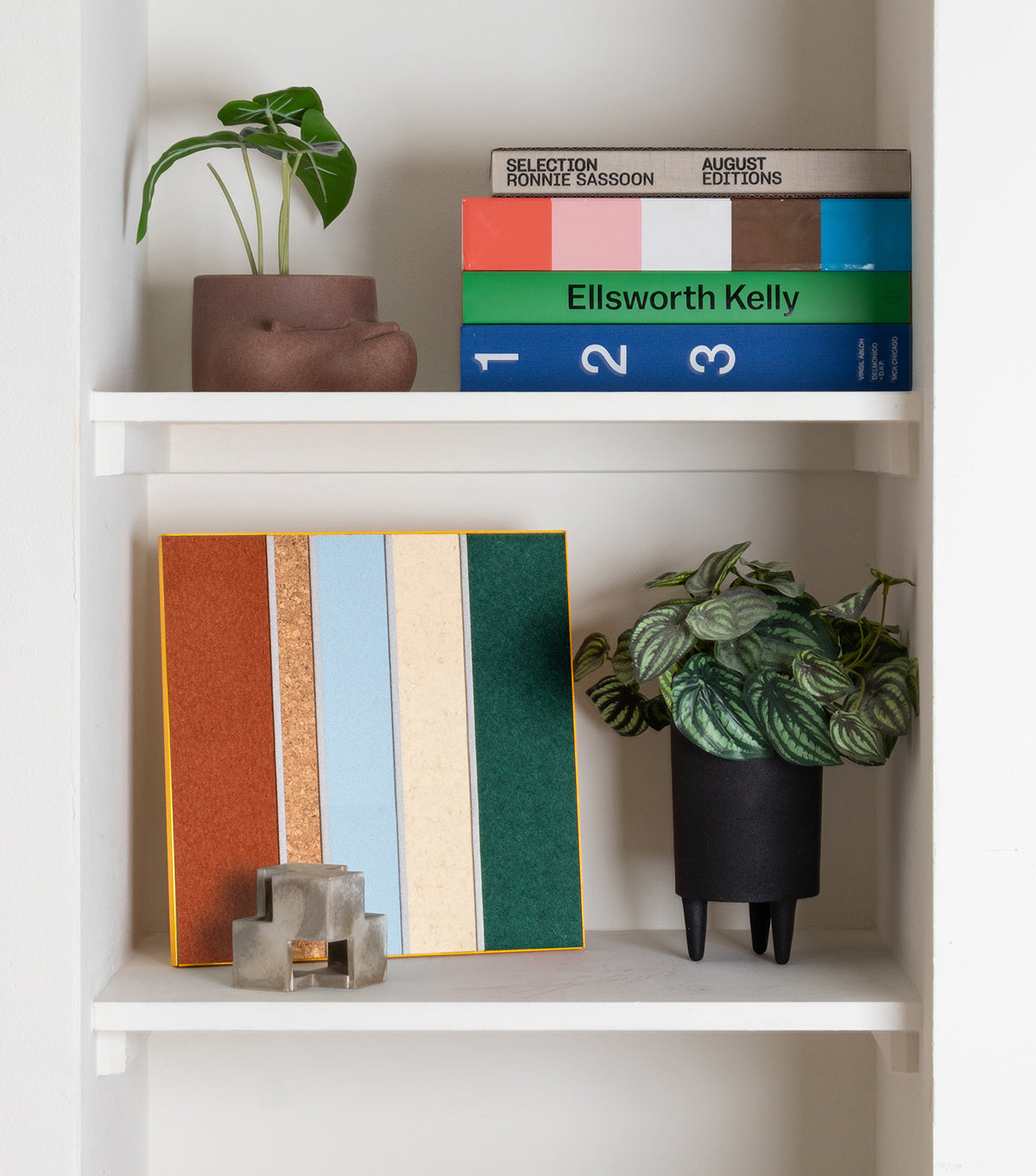 A shelf with books and a plant on the top shelf. On the shelf below is another plant and a wall art piece with strong vertical lines in orange, blue, tan, green and a light brown cork.