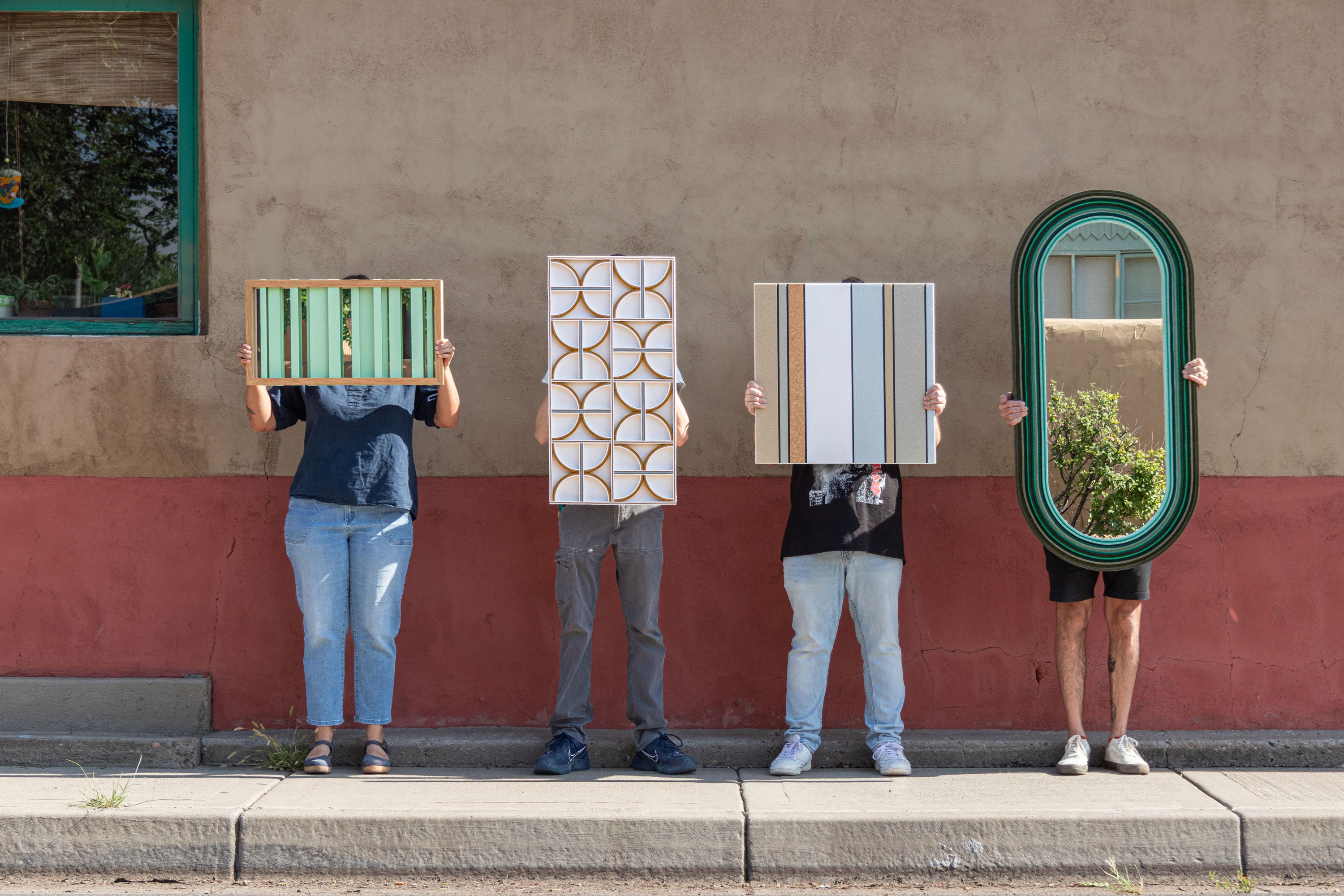 Four people holding up different mirrors and wall art pieces against a two-tone building wall.