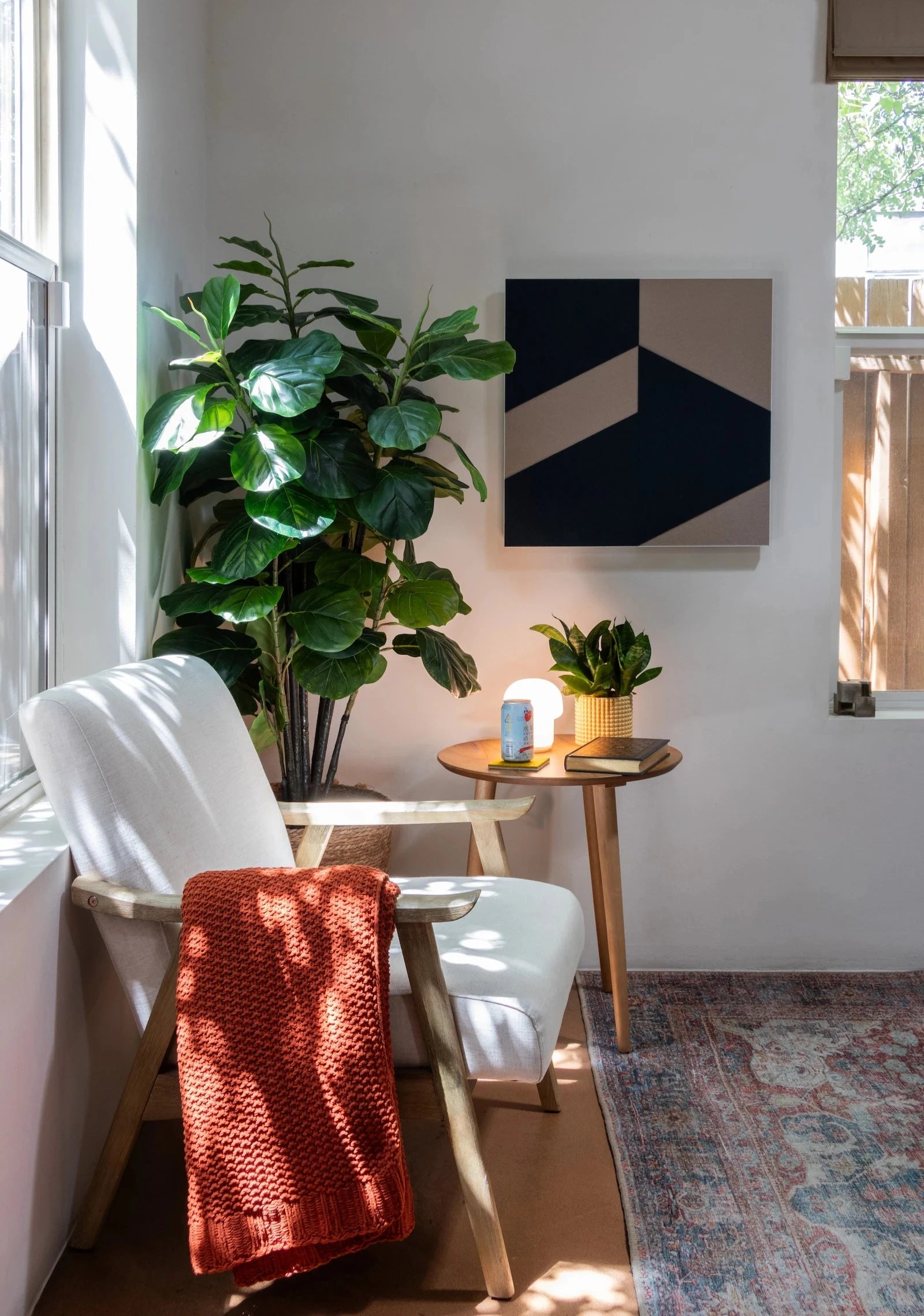 A bright corner of a living room space. A blue and tan felt wall art panel hangs on the wall with bold geometric shapes. Ar left is a large plant, a small side table and a white chair with a orange knit blanket #color_Blue/tan B