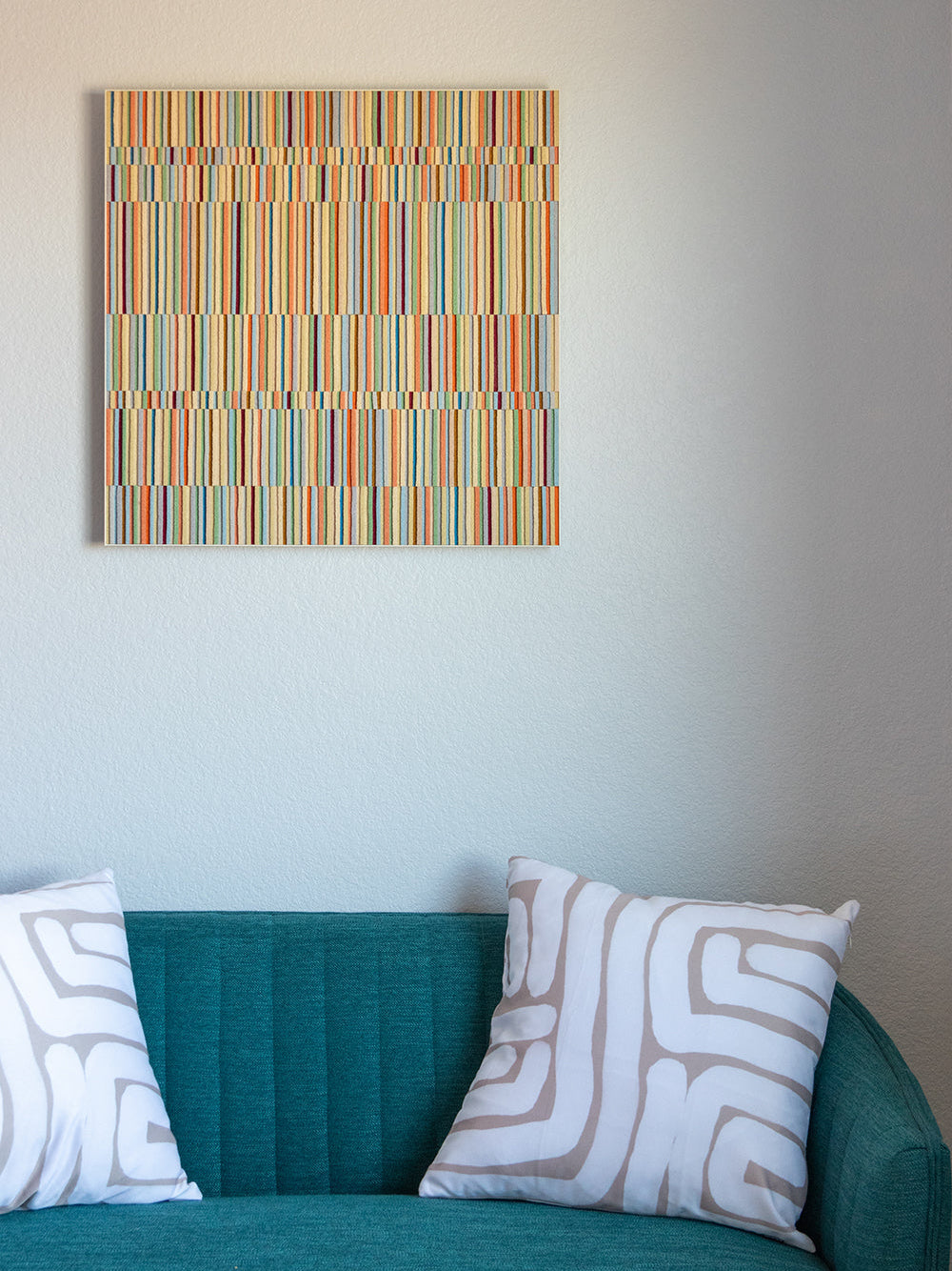 A square wall art piece hangs on a white wall. The piece is rows of alternating height wool felt strips in pastel colors. A teal loveseat with two tan and white pillows sits below the piece.