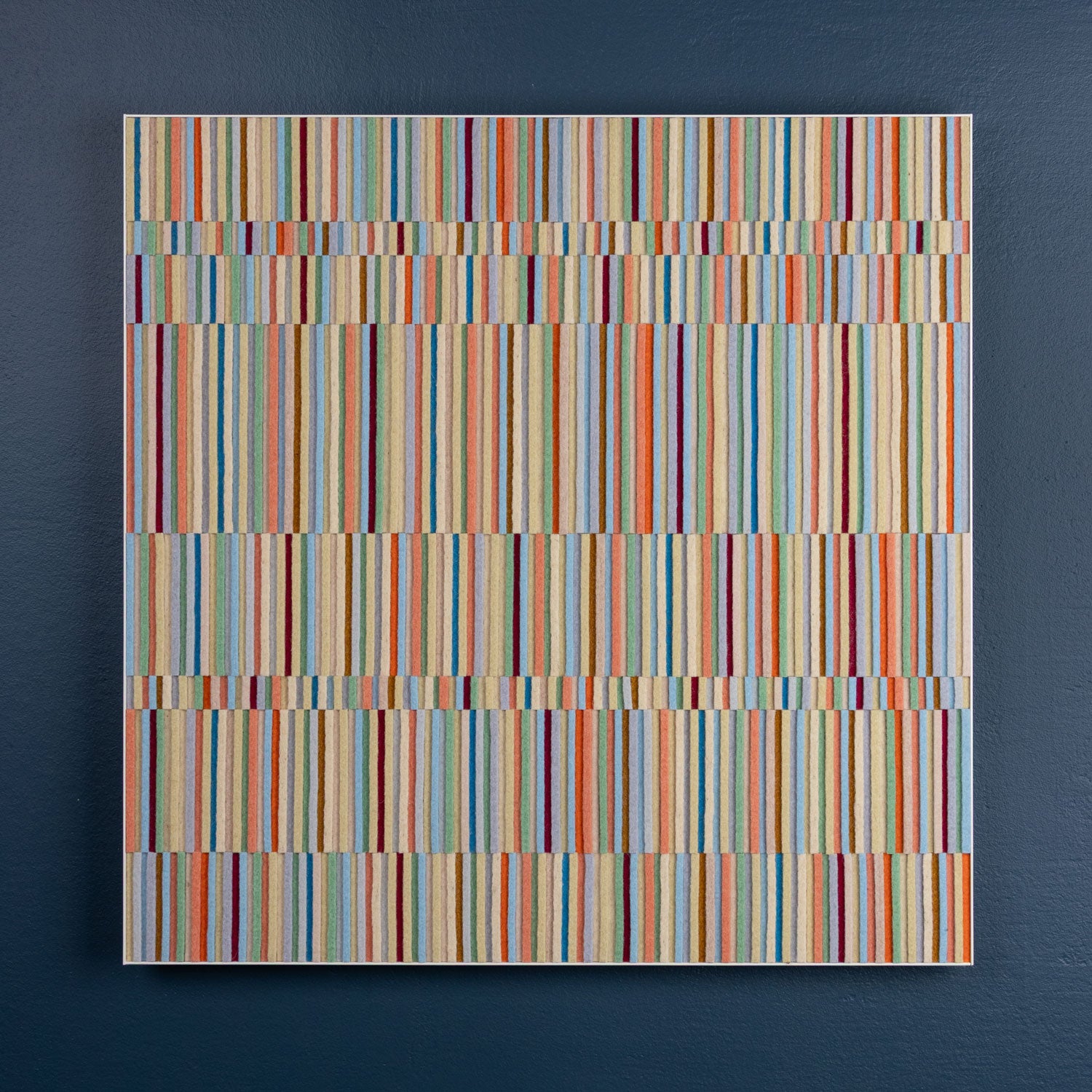A square wall art piece hangs on a dark blue wall. The piece is rows of alternating height wool felt strips in pastel colors in a white metal frame. #color_pastels