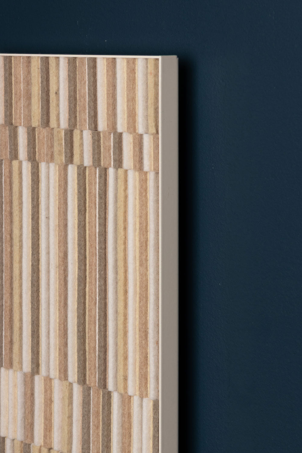 The corner detail of a ectangular wall panel with a pattern of vertical strips of felt in varying heights in ecru and beige tones, set against a dark background.