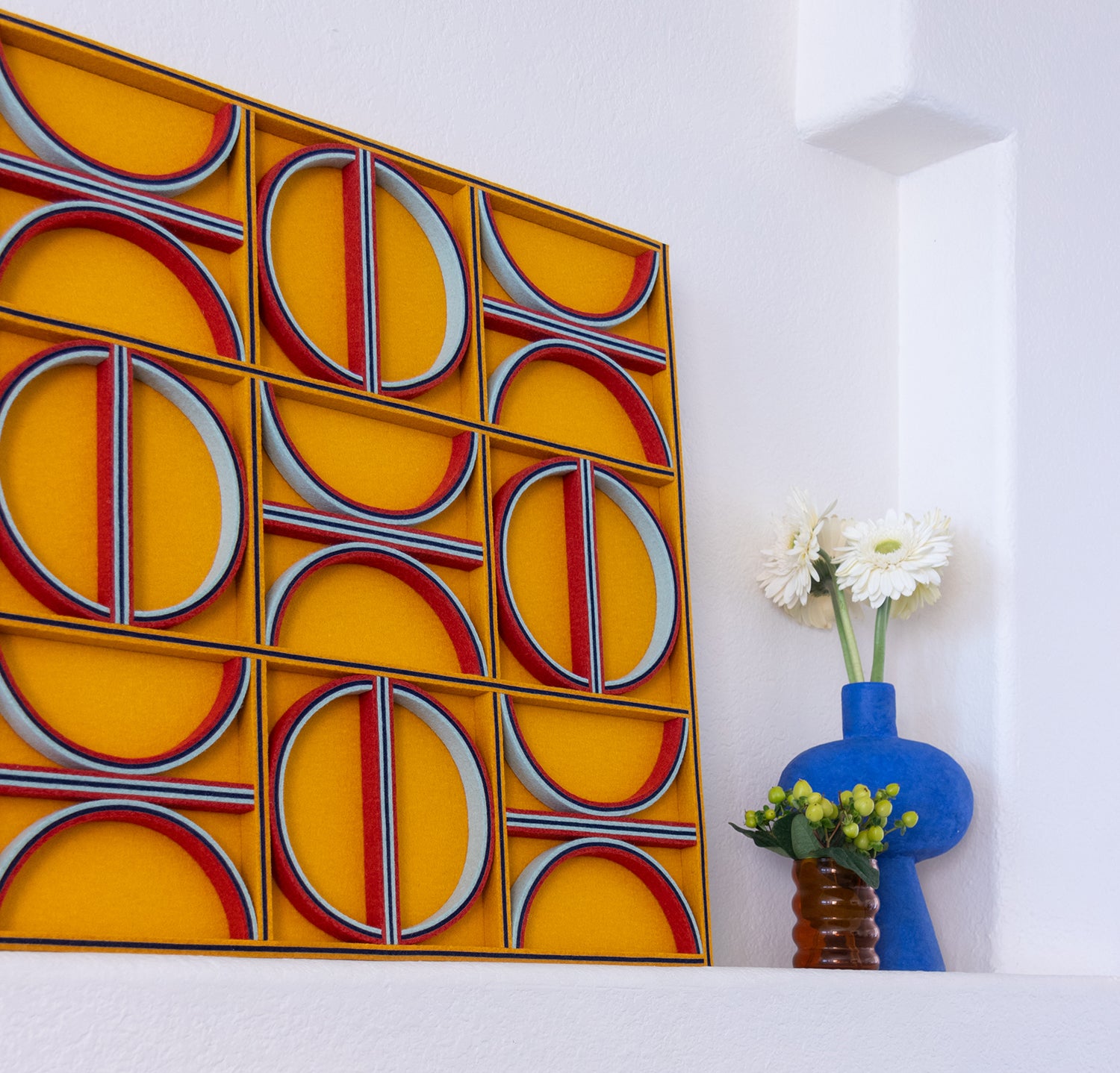 A square felt wall art panel leans on white mantel. The felt panel is a design of straight lines and half circles in blues and red on a mustard yellow background. Two vases in blue and amber hold flowers to the right of the panel.