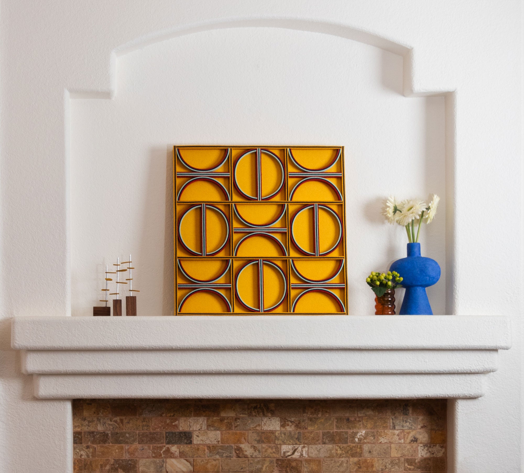 A square felt wall art panel leans on white mantel above a brick fireplace. The felt panel is a design of straight lines and half circles in blues and red on a mustard yellow background.