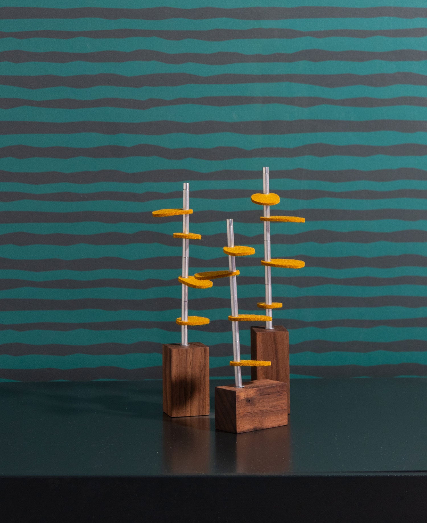 Three walnut wood rectangle bases with metal stems and ochre colored felt leaves sit on a black surface with a wiggly horizontal striped wall paper in gray and green. #color_Walnut/ Ochre