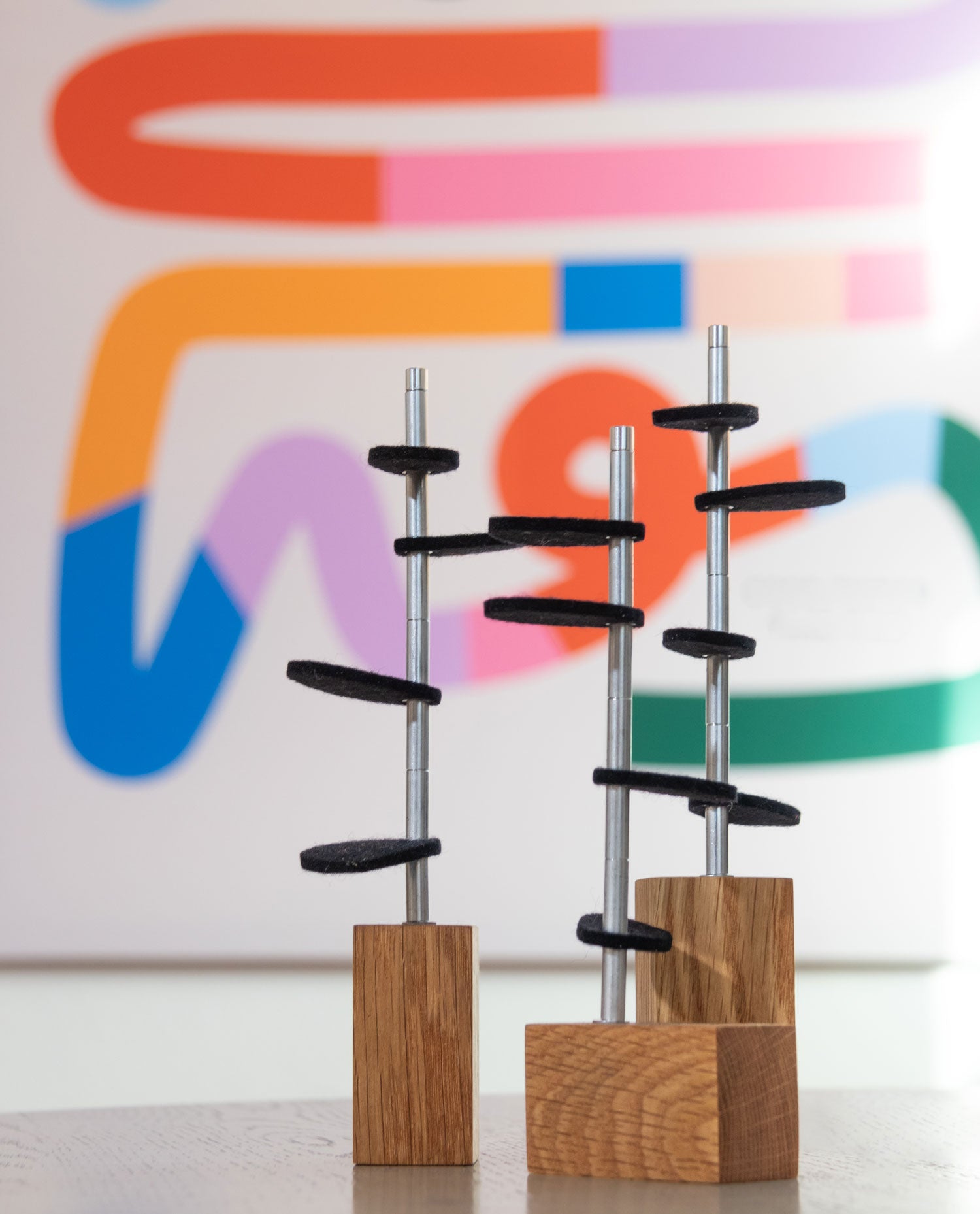 Three white oak wood rectangular bases with metal stems and black wool felt leaves sit on a surface in front of colorful squiggle line art. #color_White Oak/ Black
