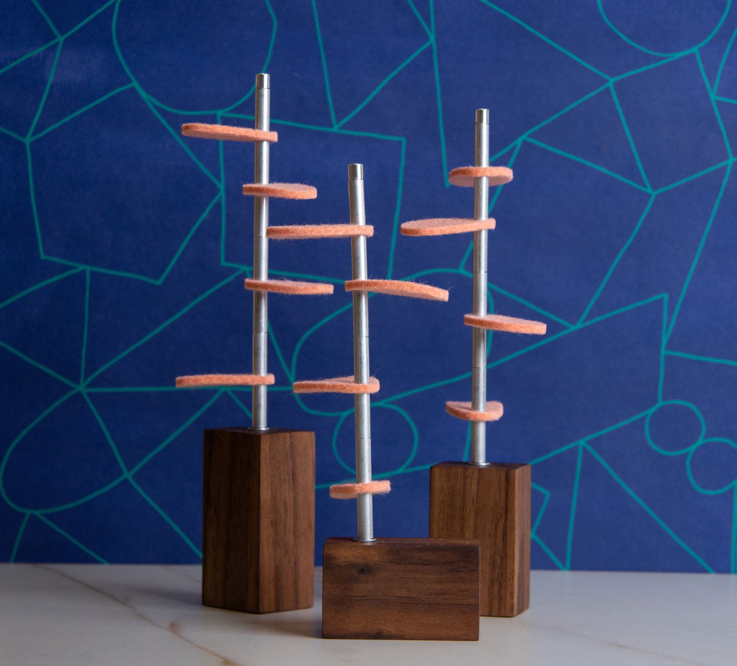 Three walnut wooden blocks with metal stems and coral colored wool felt leaves sit in front of a two tone blue wallpaper #color_walnut/ coral