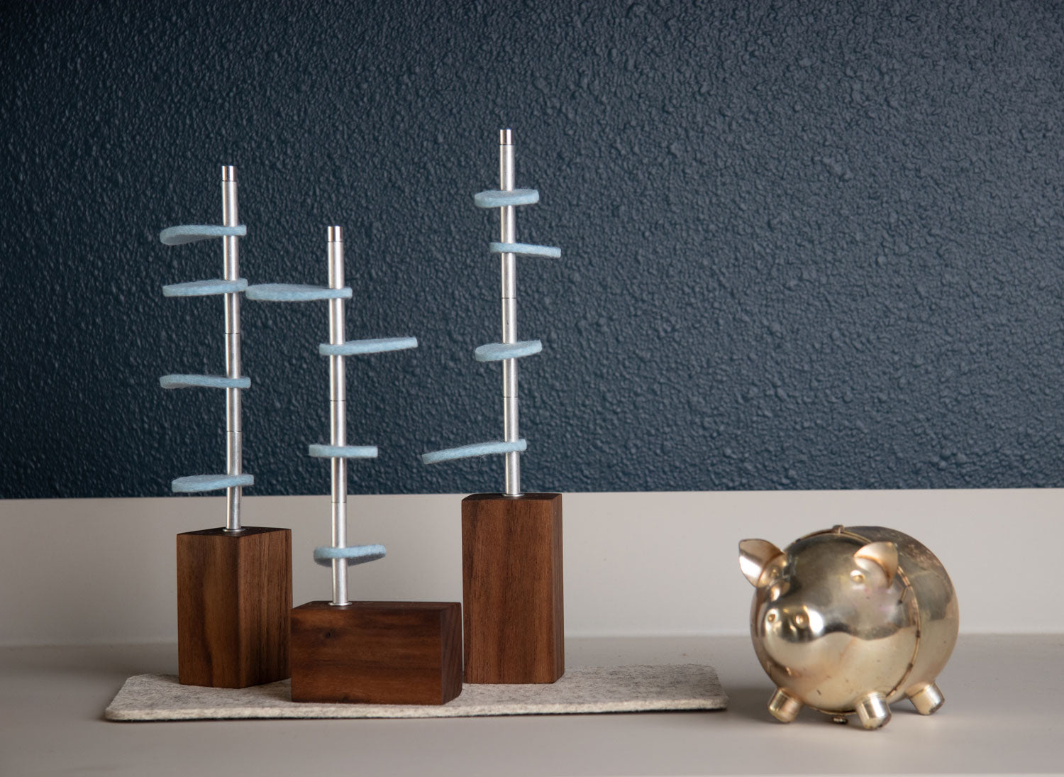 Three walnut wood rectangular bases with metal stems and ice blue wool felt leaves on a gray felt mat. They are on a gray shelf with a small metal piggy bank. #color_Walnut/ ice