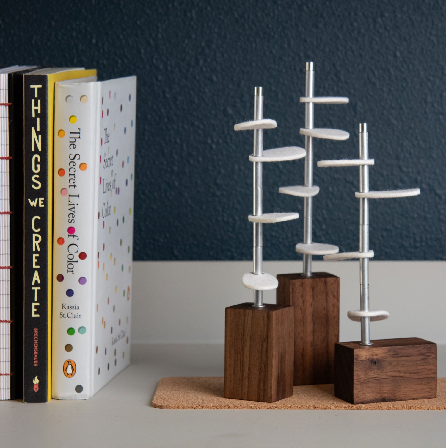 Three walnut wood bases with metal stems and white wool felt leaves sit on a cork mat. Next to the sculptural objects are books. #color_Walnut/ off white