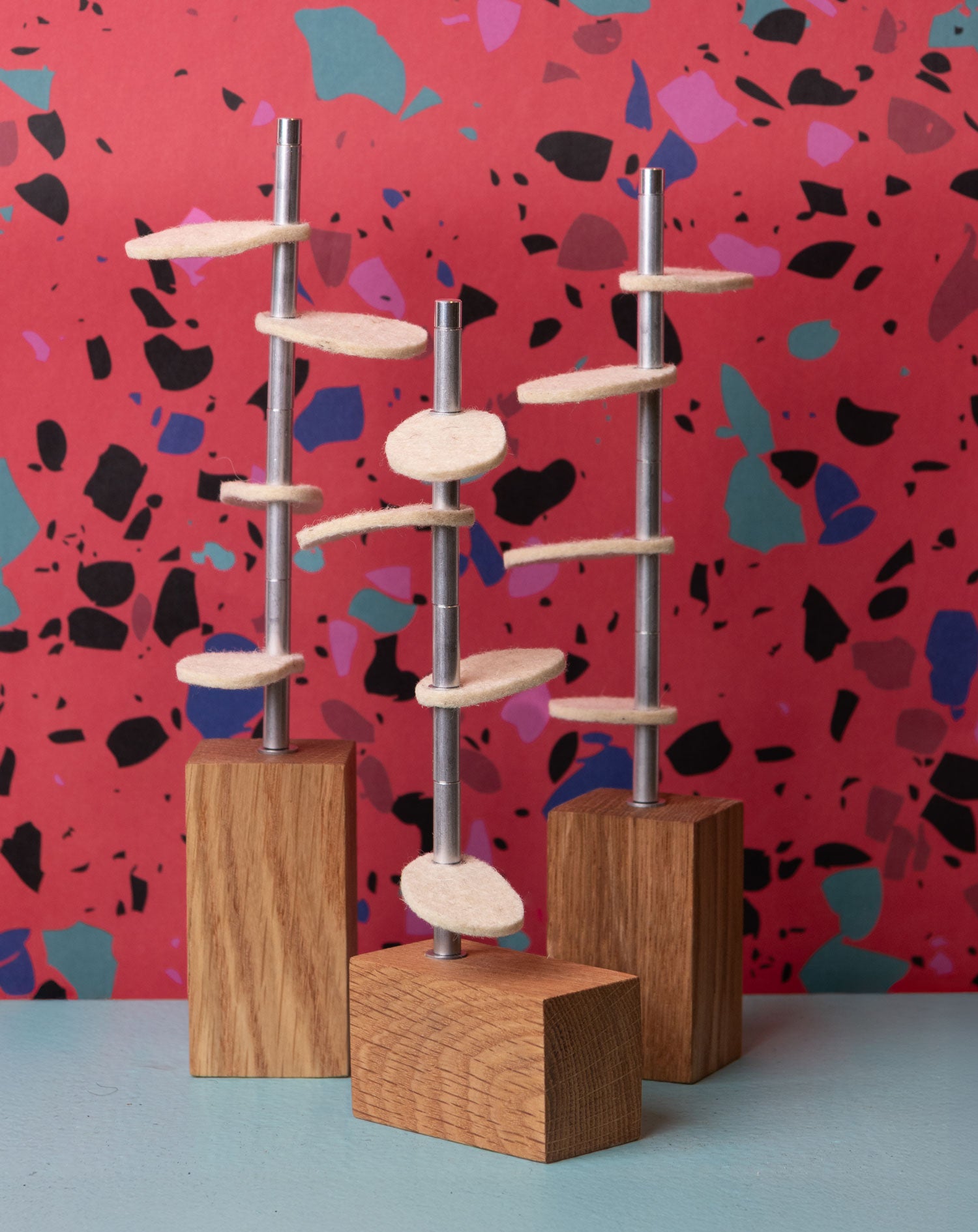 Three white oak wood rectangular bases with metal stems and cream colored wool felt leaves sit on a light blue surface in front of a red terrazzo wallpaper. #color_White oak/ raw white