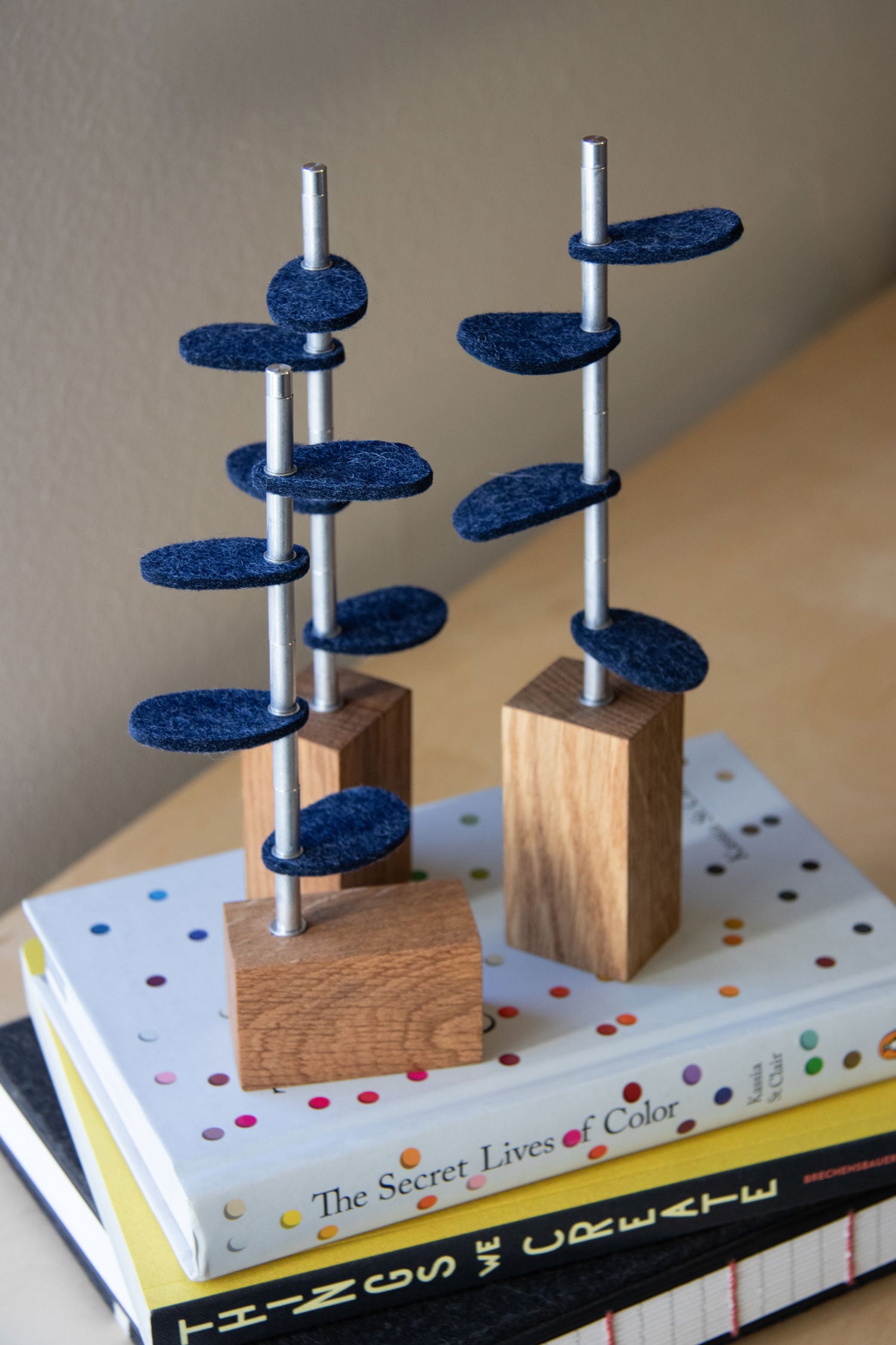 Three white oak bases with metal stems and blue heathered wool felt leaves sit on a pile of books. #color_White Oak/ Indigo