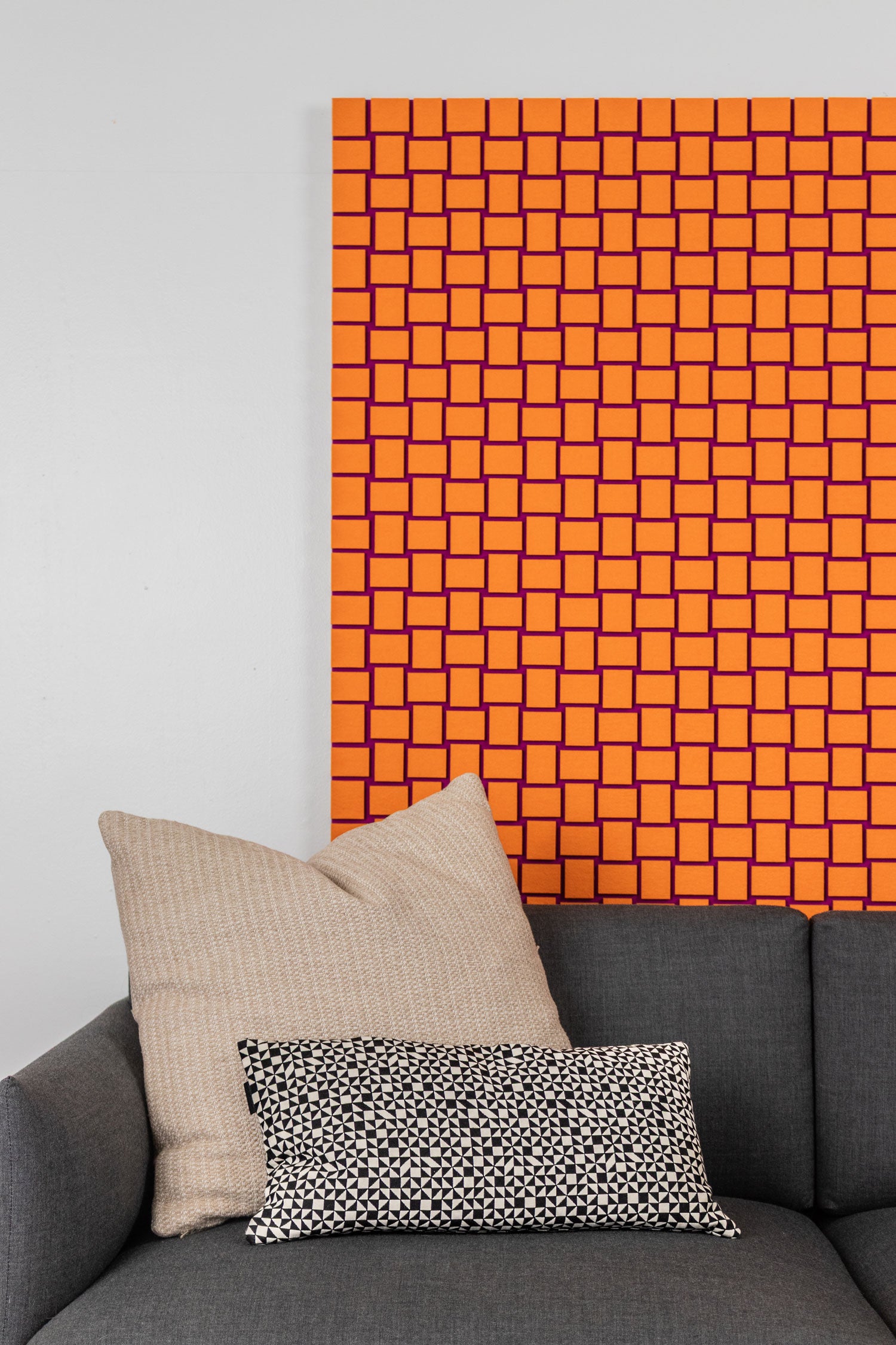 Gray sofa with beige and patterned cushions against an orange felt wall panel with a repeating square pattern