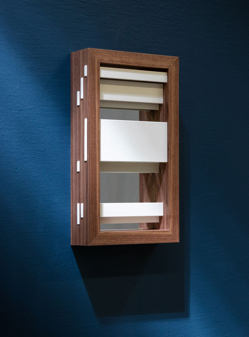 A rectangular mirror with a solid wood frame, featuring white horizontal powder-coated louvers in various widths, seen on an angle on a blue wall.