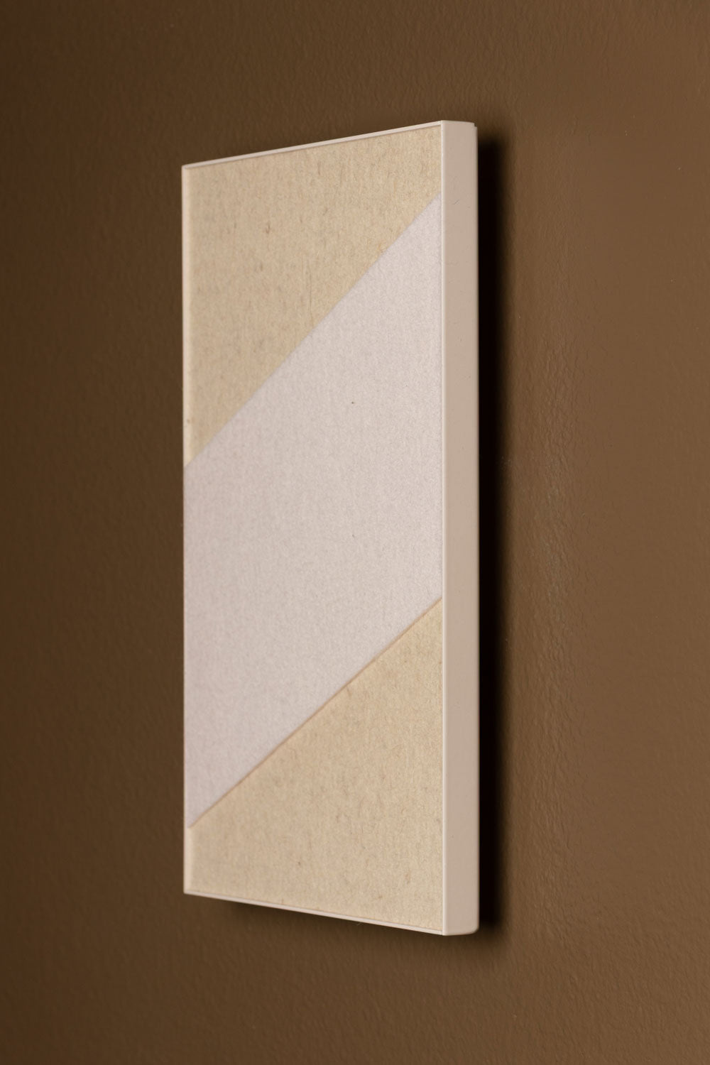 A rectangular wall panel with a geometric pattern consisting of angular fields of wool felt in varying shades of beige and white, with a powder-coated frame on a brown wall.