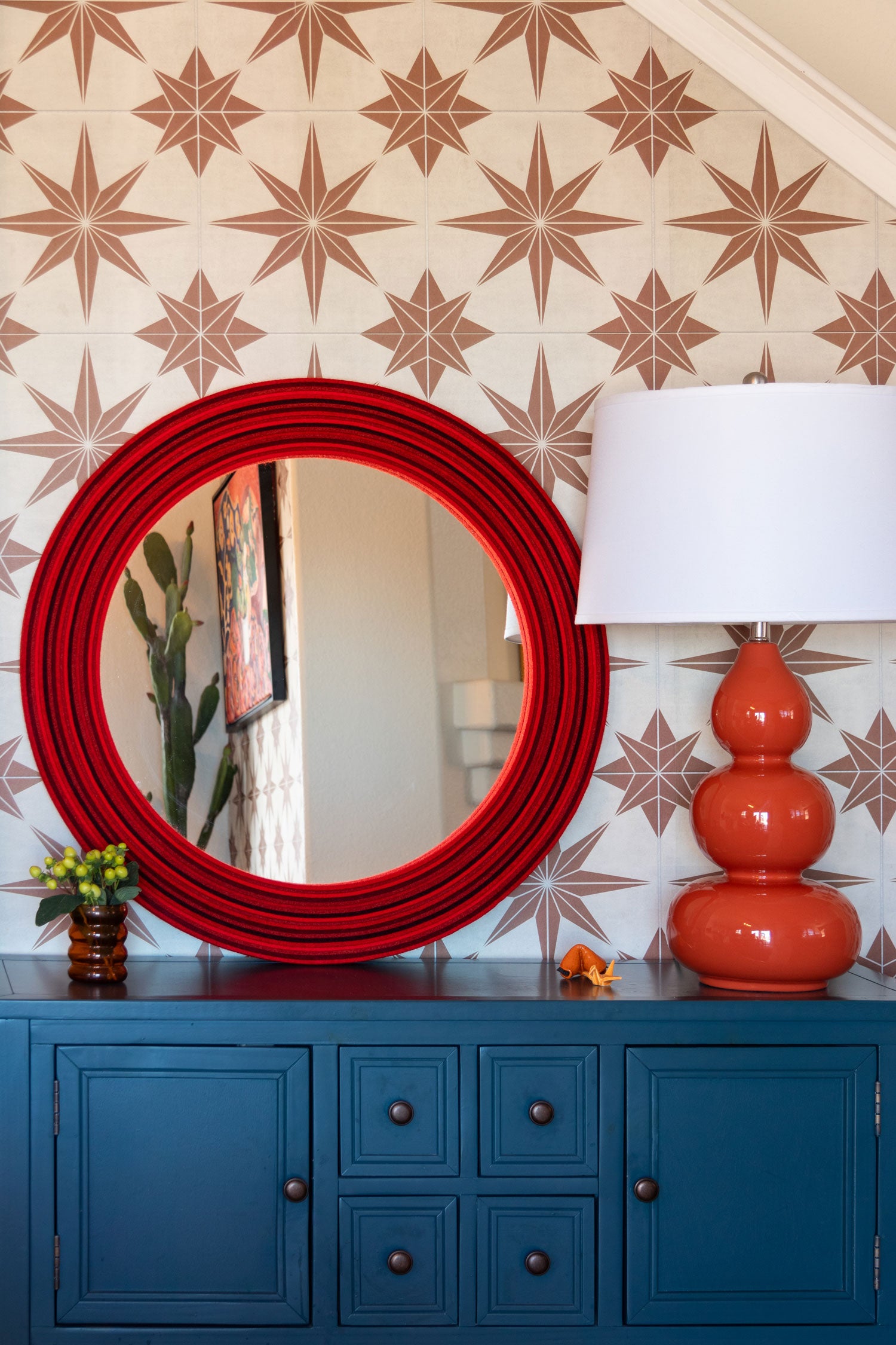 A large round mirror framed with multi-tone red felt leans on a patterned wall above a blue sideboard. The surface of thr sideboard also has a small vase of flowers, an orange lamp, and some knick knacks.
#color_multi-color reds