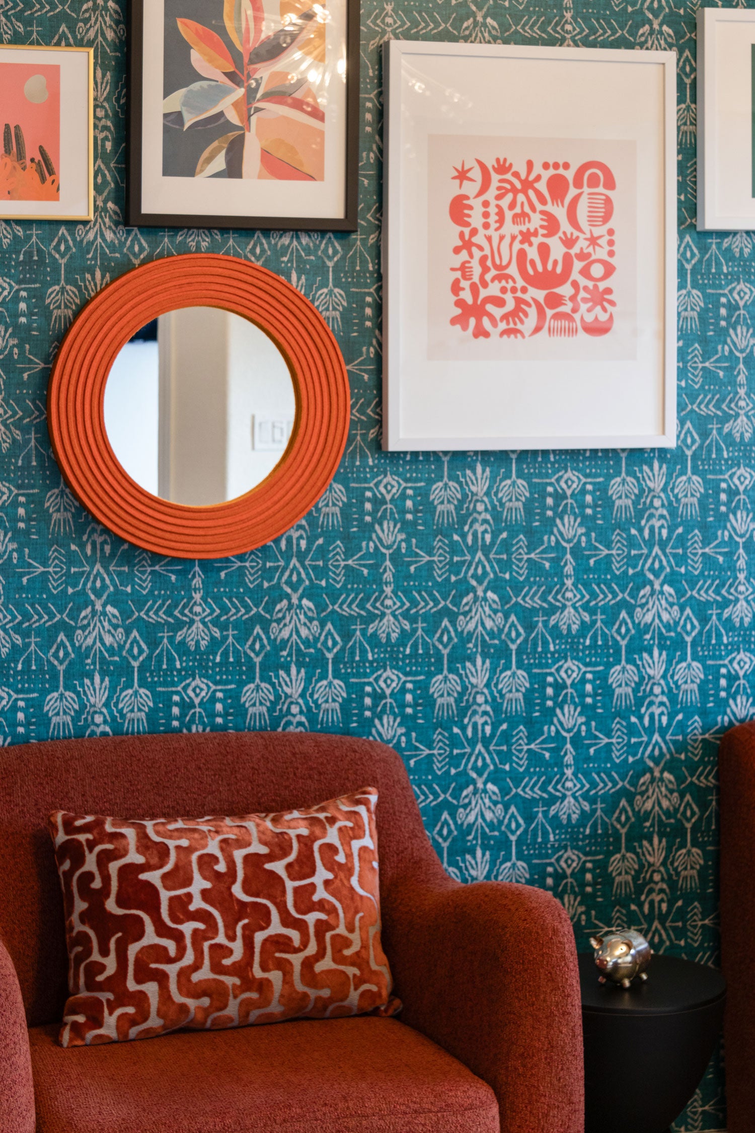 A small round mirror framed in orange felt hangs on a wall of blue patterned wall paper. There are other frames artworks on the wall and a red cushioned chair below the pieces on the wall.