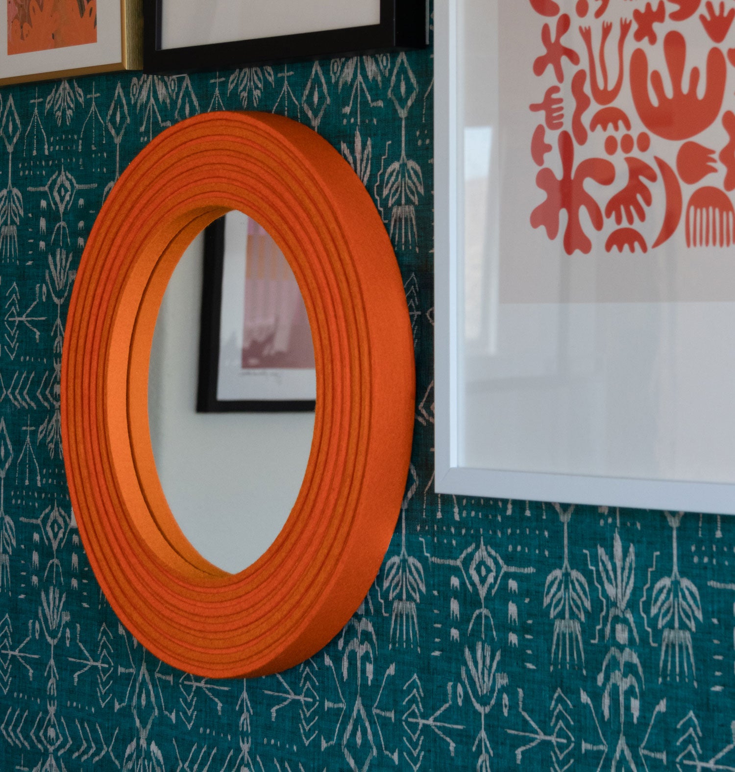 A closeup of a small round wall mirror framed with orange felt on edge. It hangs on blue and white patterned wallpaper with other framed art pieces.