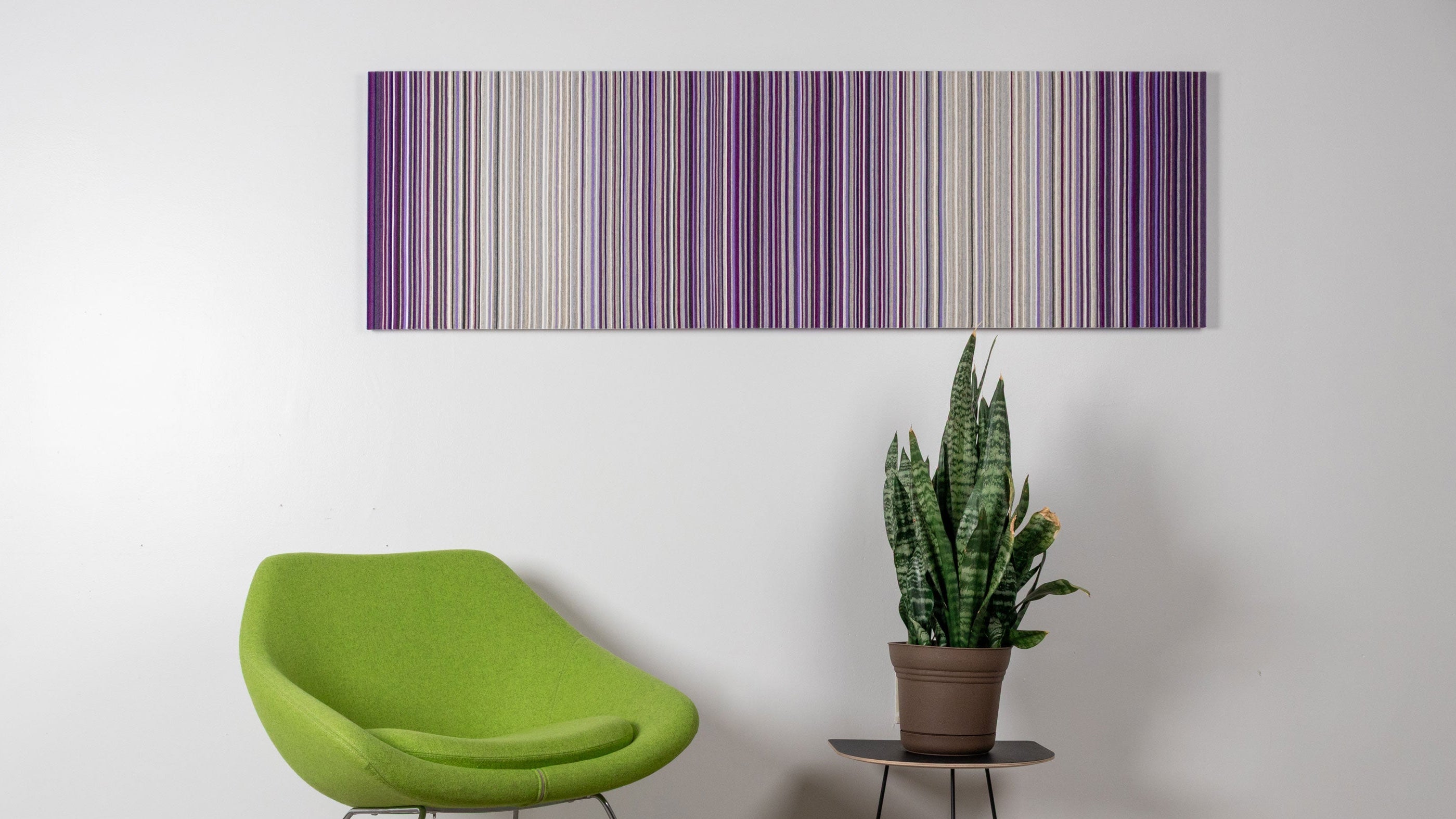 Green modern chair and potted plant against a white wall with a purple and gray wool felt wall art piece with very thin vertical stripes.