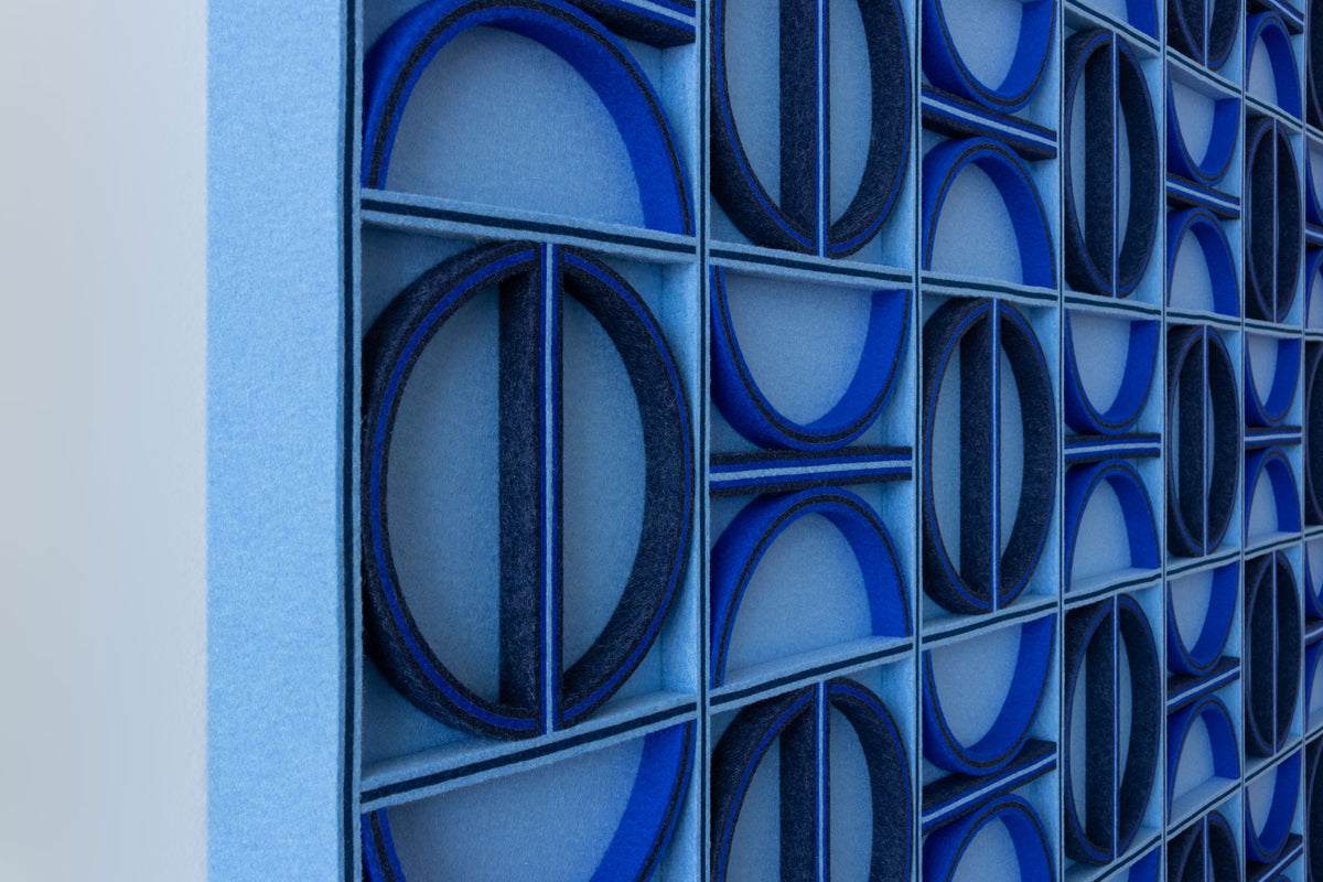 A close-up angled shot of a multi-blue-tone wool felt geometric wall panel with repeating circles and half circles in a grid.