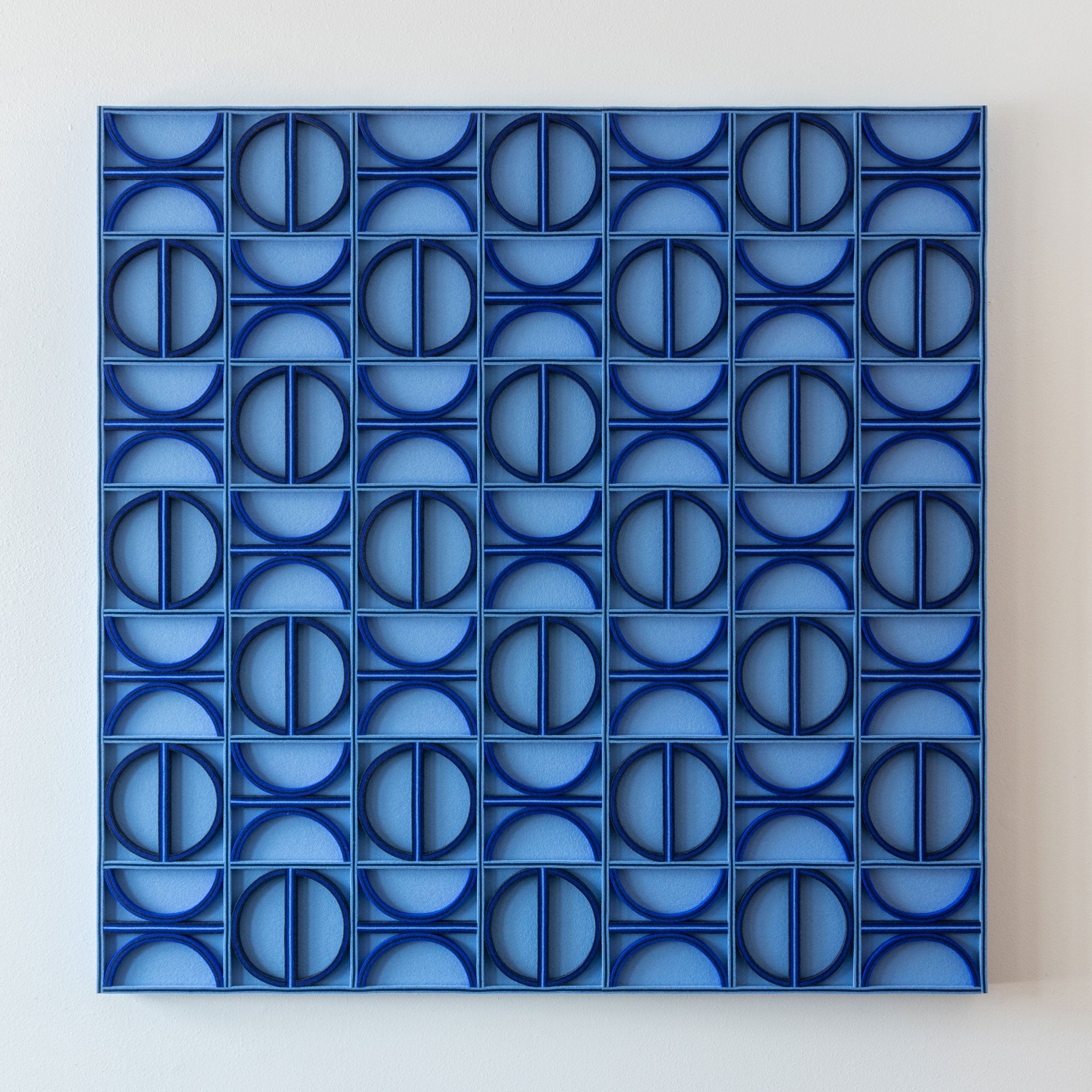 A square blue wool felt geometric wall art piece featuring a repeating pattern of interlocking circular and square forms, mounted against a plain light wall.
#color_sky blue