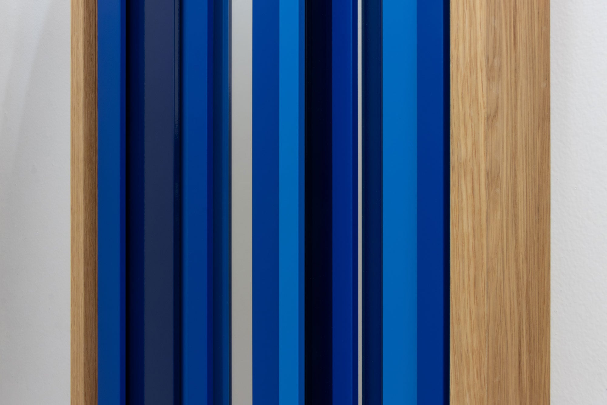 Abstract mirror featuring vertical stripes in various shades of blue, navy, and a light wood grain frame mounted on a white wall.