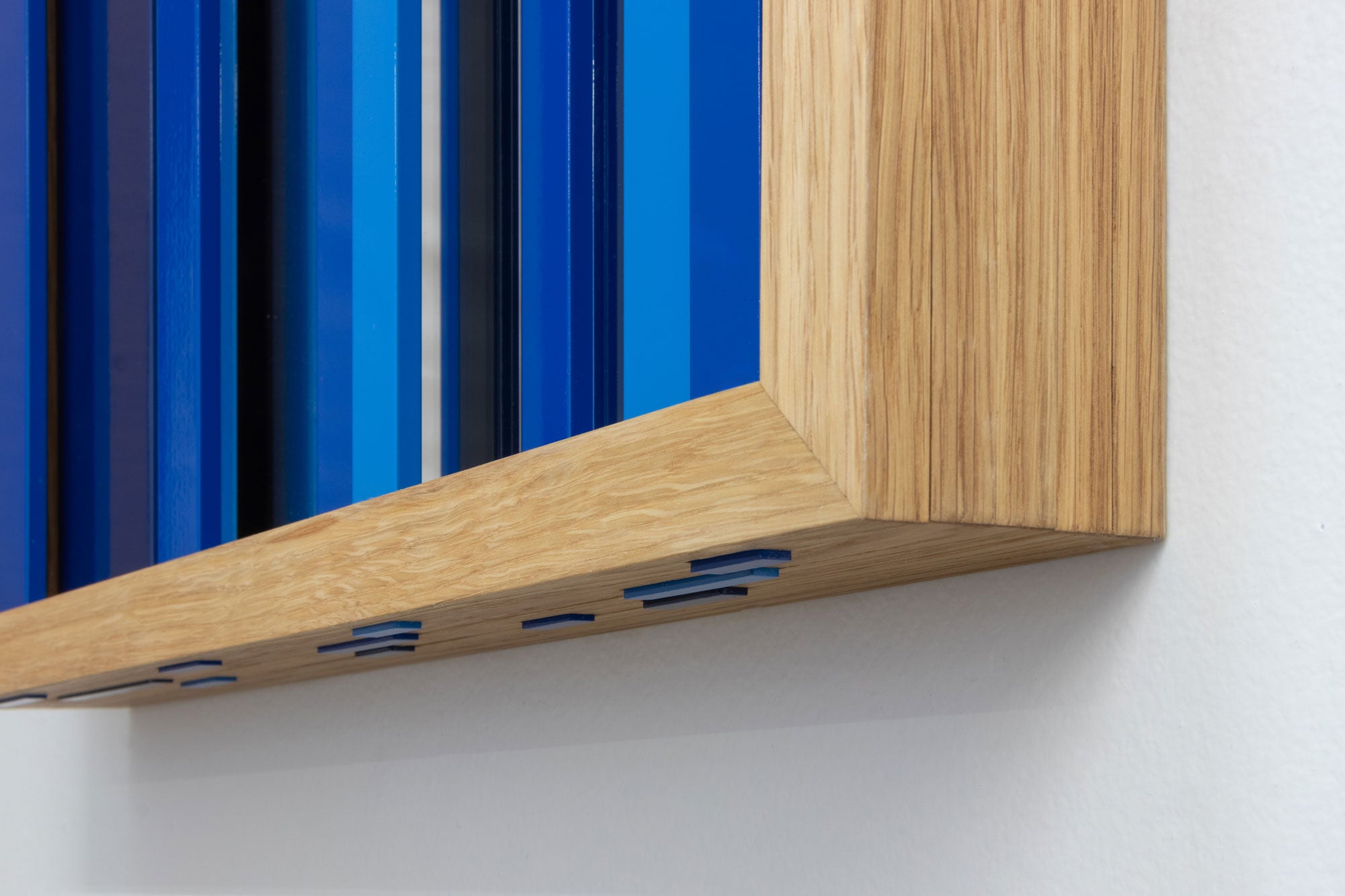 Close-up of a modern wall-mounted art piece featuring vertical blue and navy panels framed by a light oak wood border, mounted on a white wall.
