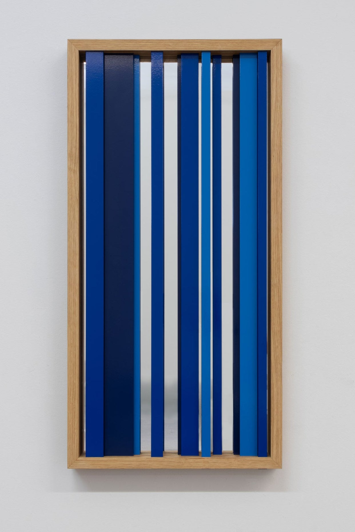 A modern decorative rectangular mirror with a white oak frame, multi-color blue louvers and a glass mirror behind the louvers, hung on a white wall.