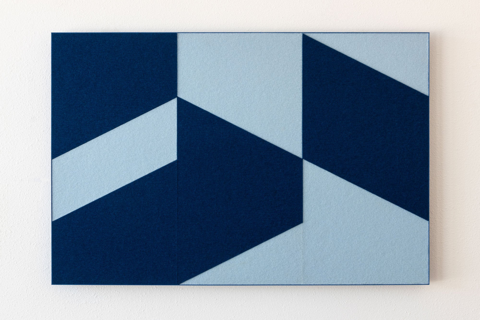A rectangle abstract wall art piece with a geometric pattern of dark navy blue and light blue shapes, mounted on a plain off-white wall. #color_Two tone blue
