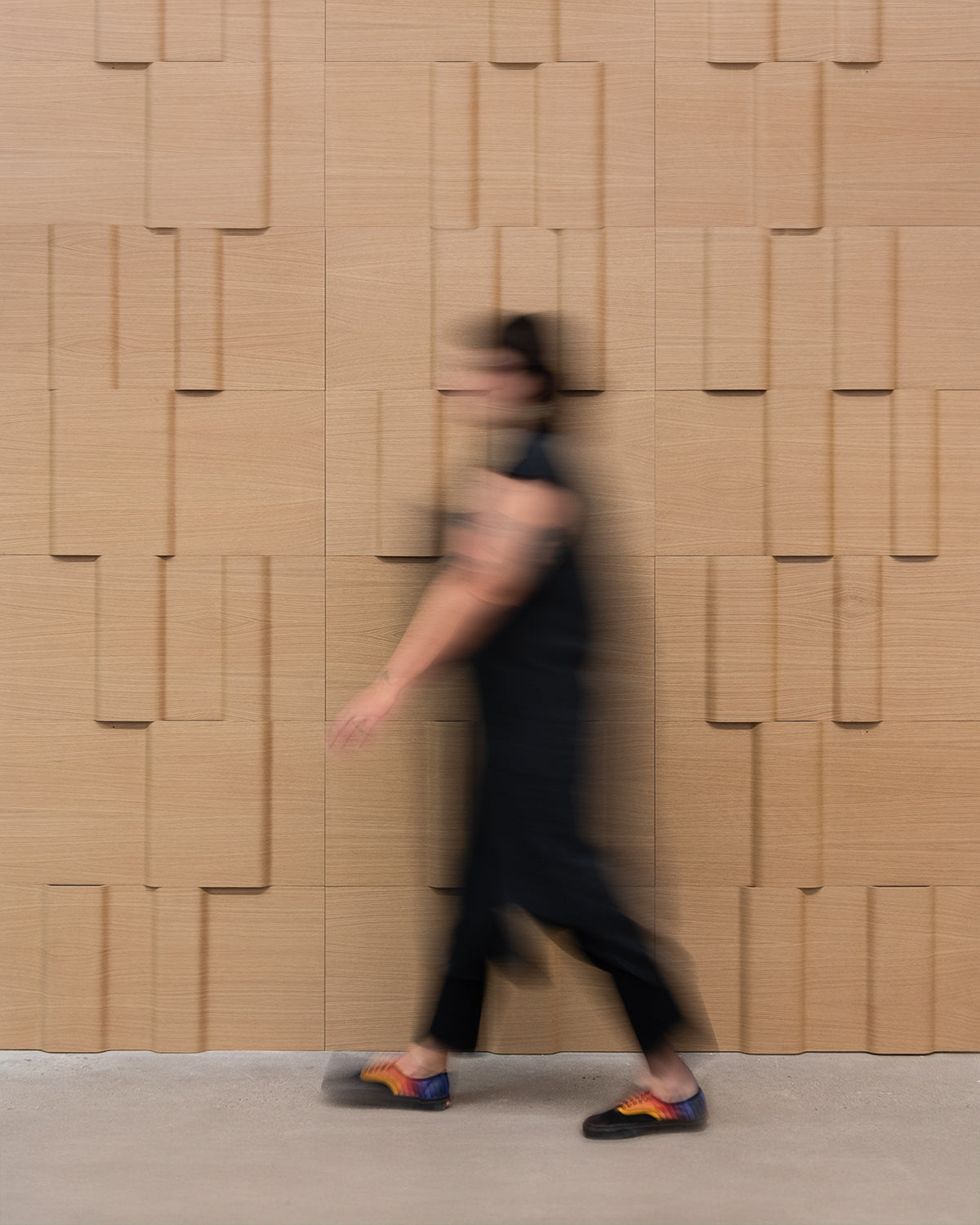 A blurred figure in black walks in front of a white oak dimensional wood wall.